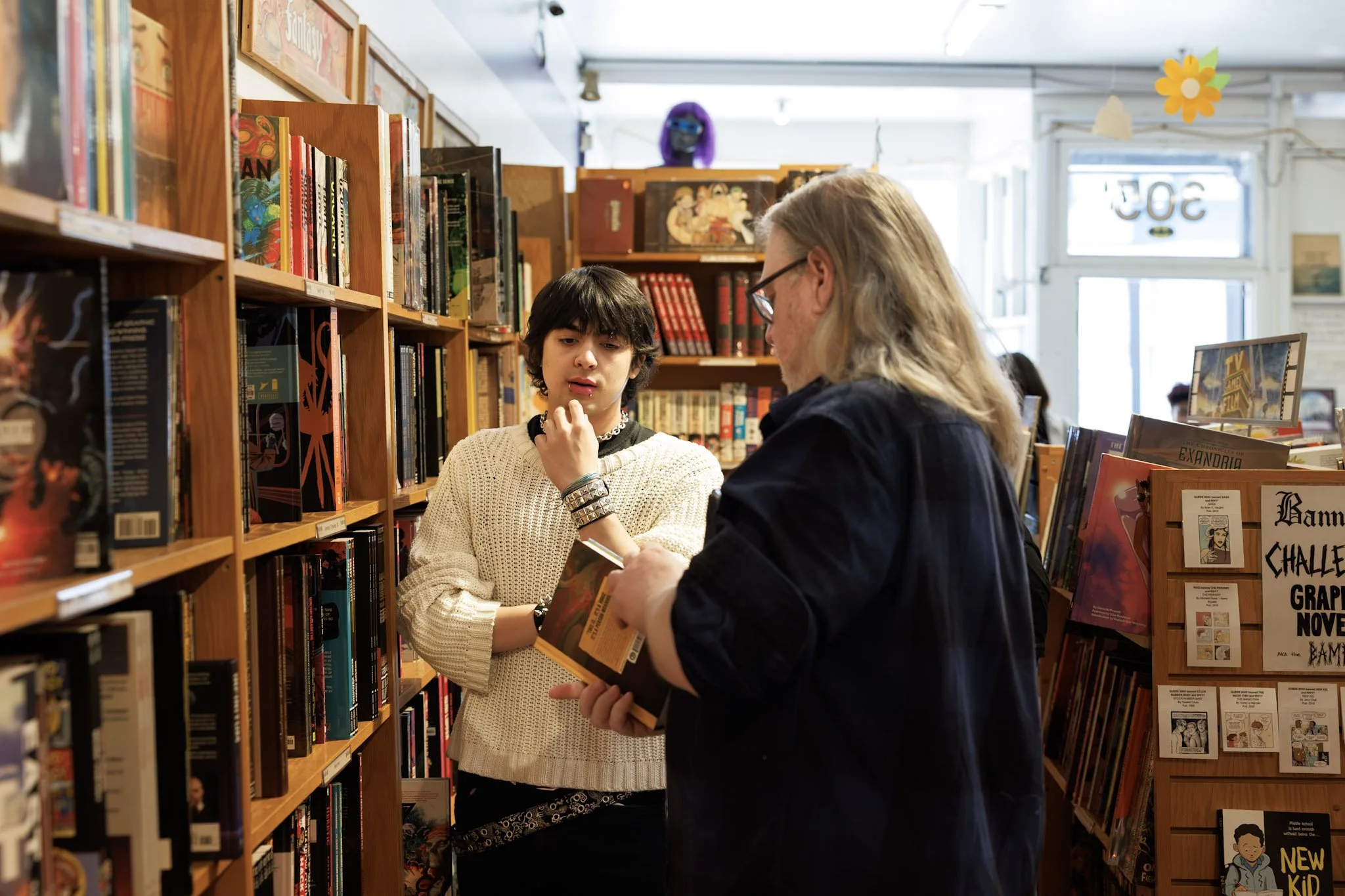 Two people inside a bookstore, one young woman with dark hair and a white sweater, and one older man with long gray hair and glasses, looking at books together among wooden shelves filled with colorful books.