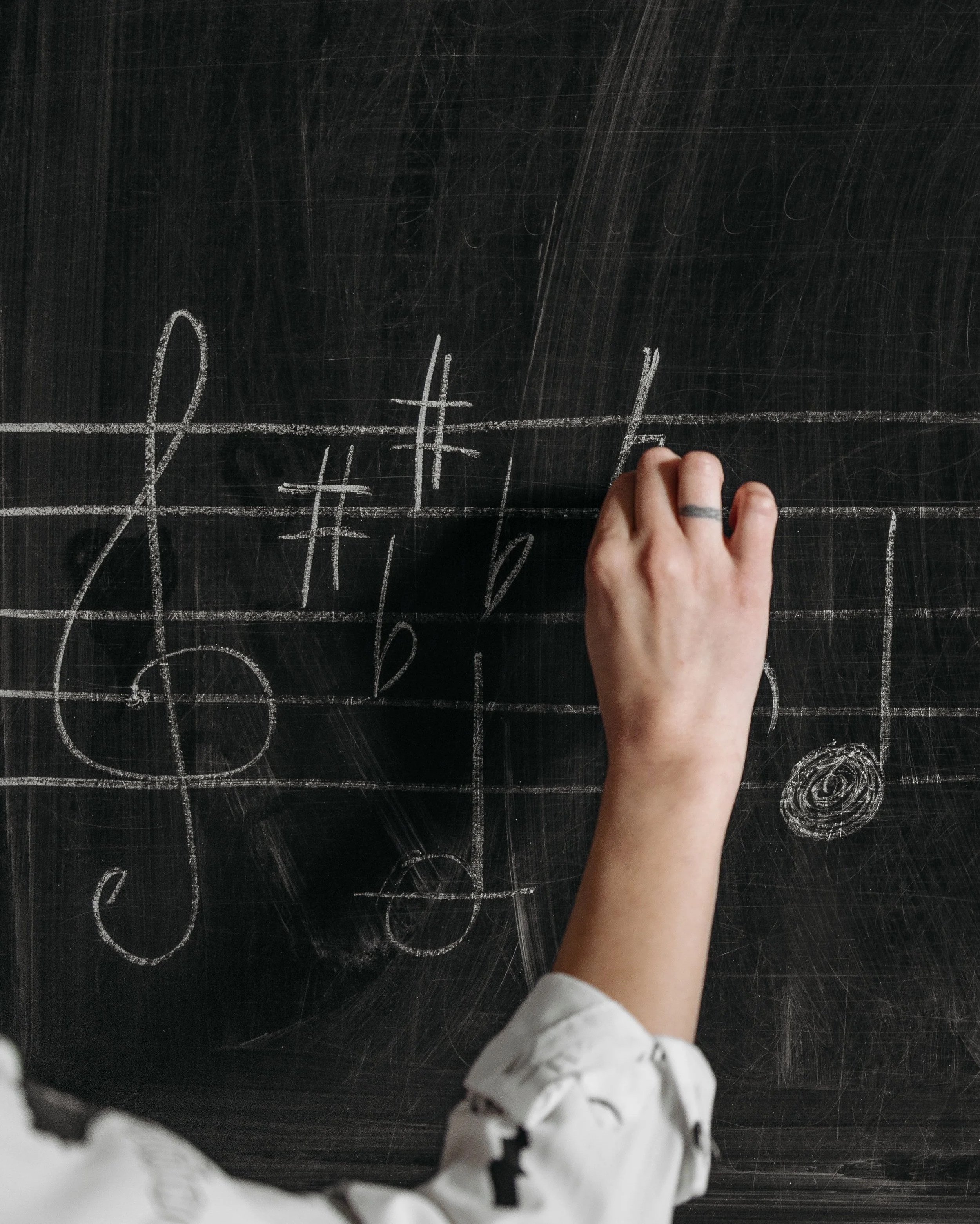 A person writing musical notes and symbols on a black chalkboard with white chalk.