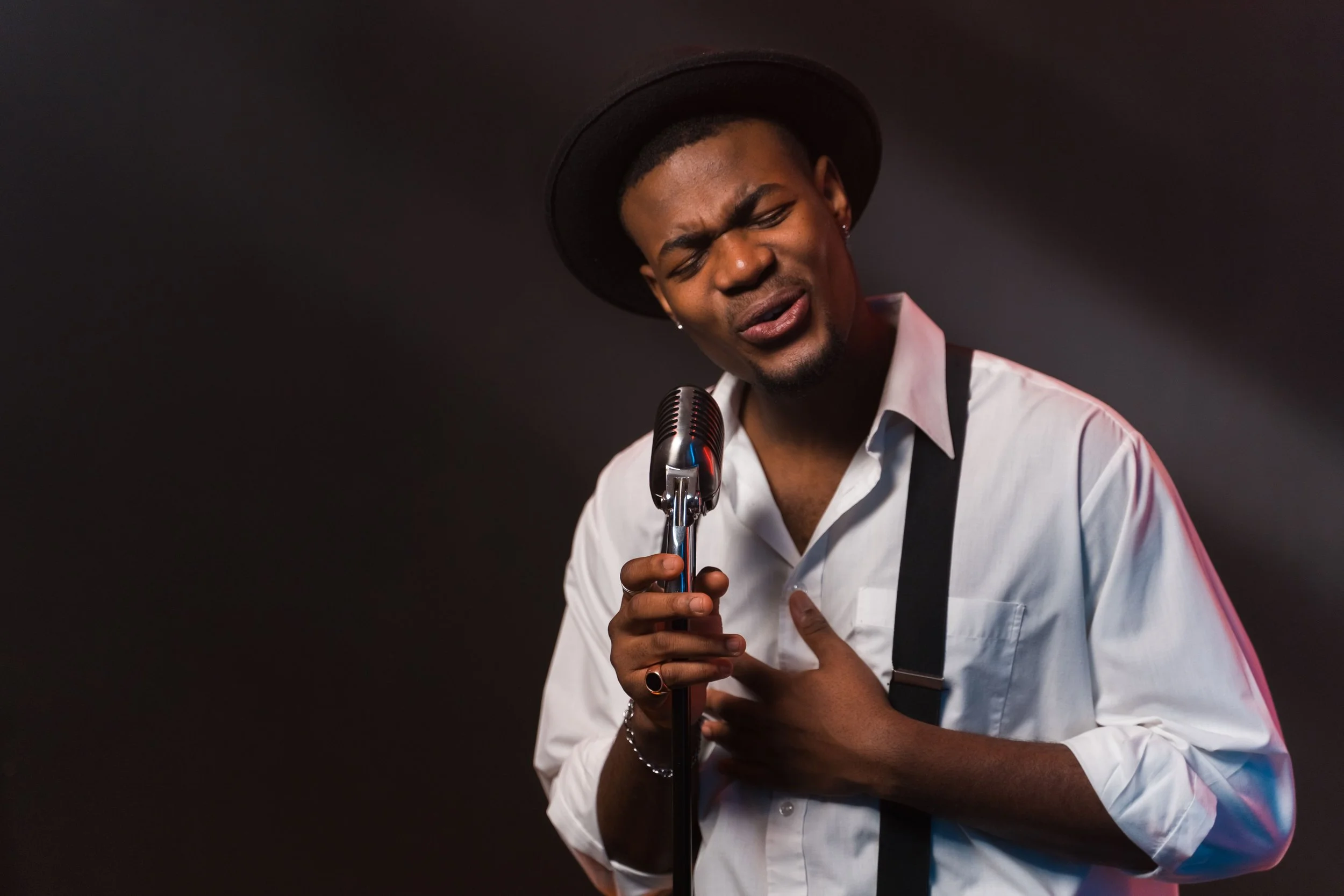 A man with dark skin and a short haircut wearing a white shirt and a black hat, singing into a retro microphone with his eyes closed and hand placed on his chest against a dark background.