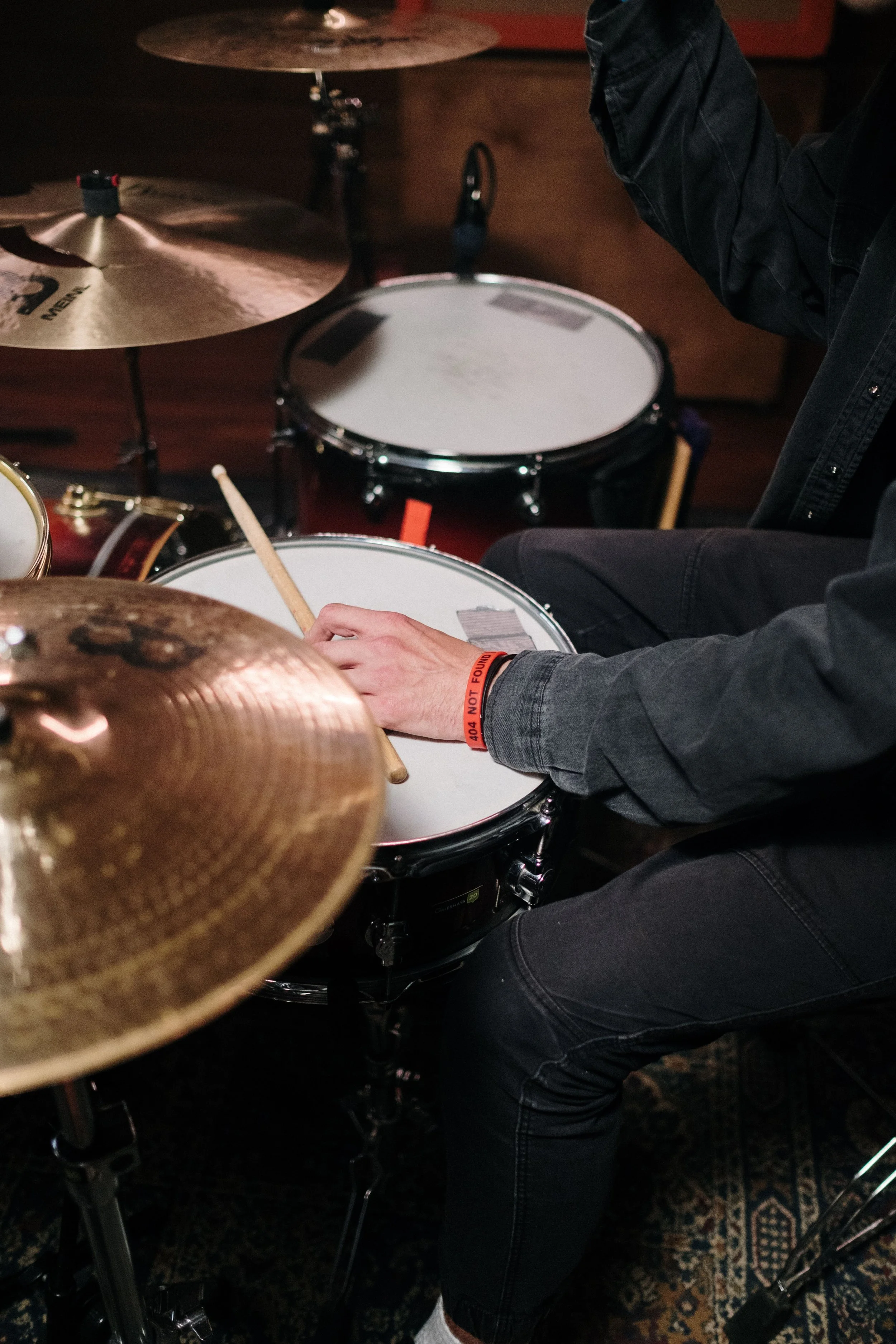 A person playing a drum set, wearing a gray shirt with an orange wristband, black pants, and holding a drumstick. The drum kit includes a snare drum, cymbals, and a bass drum, set up on a dark patterned rug.