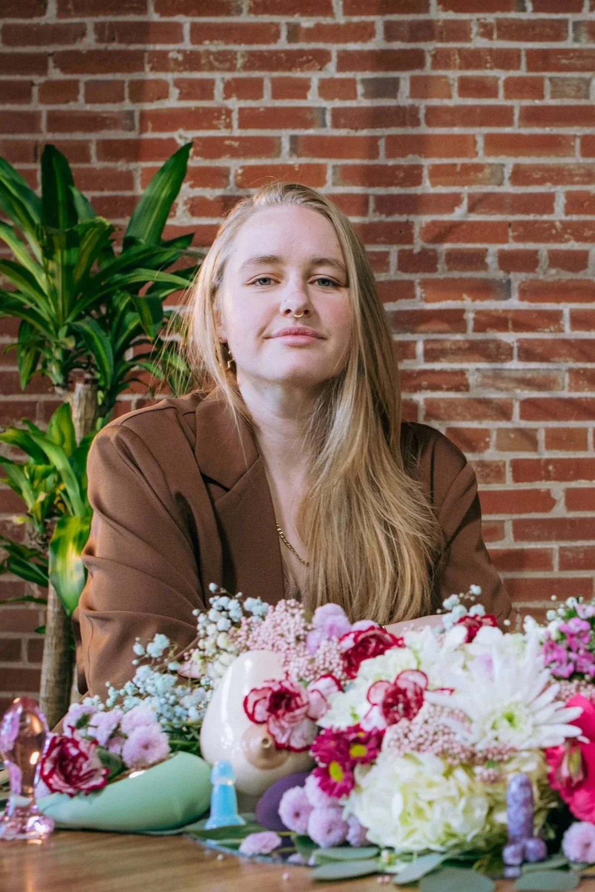 A white non-binary person with long blonde hair, wearing a brown blazer, sitting at a table with a large floral arrangement that contains sexual health paraphernalia. Behind them is a brick wall and a green plant.