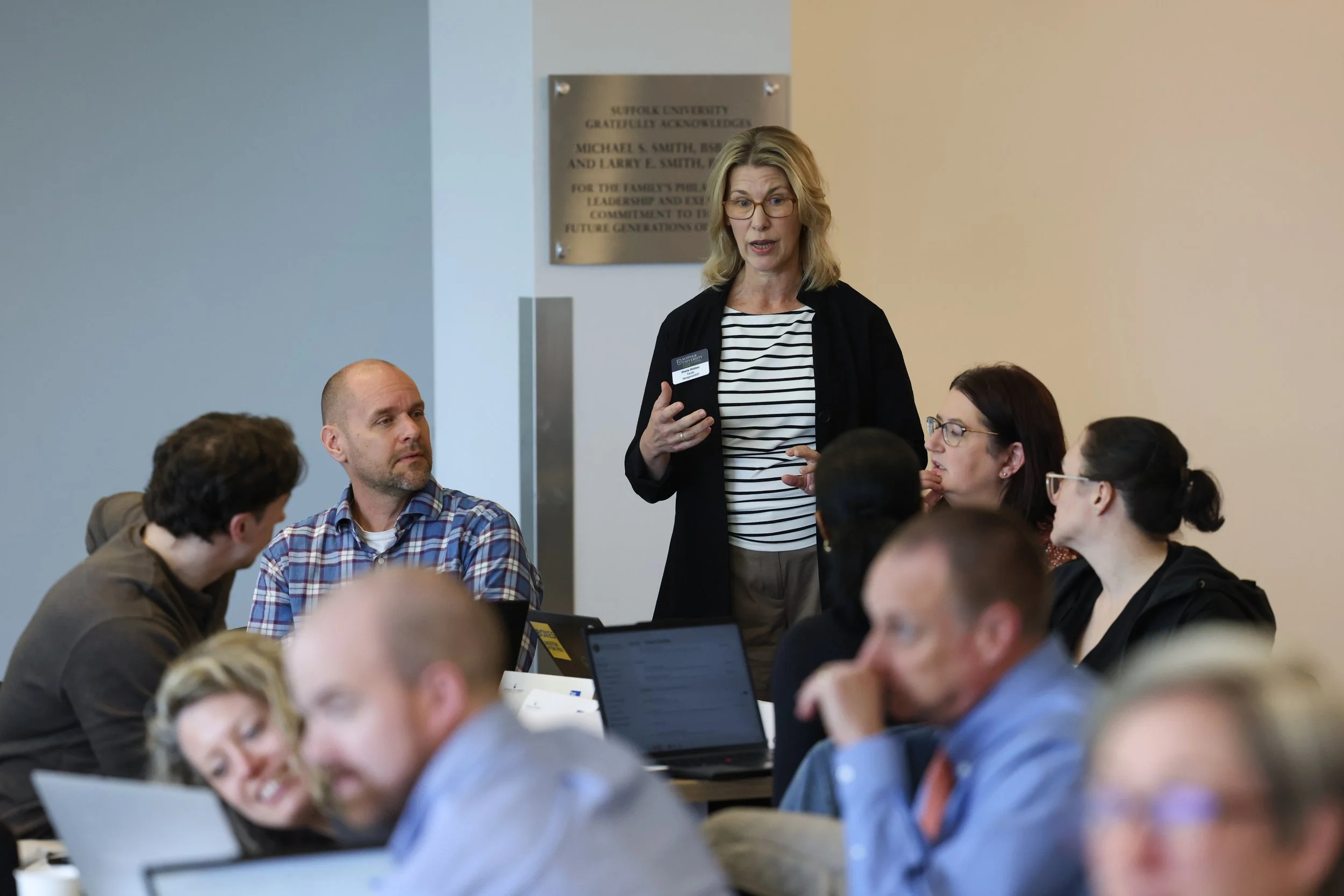 A woman with blonde hair, glasses, and a striped shirt speaking to a group of people at a conference or meeting in a room with a gray wall and a university plaque in the background.