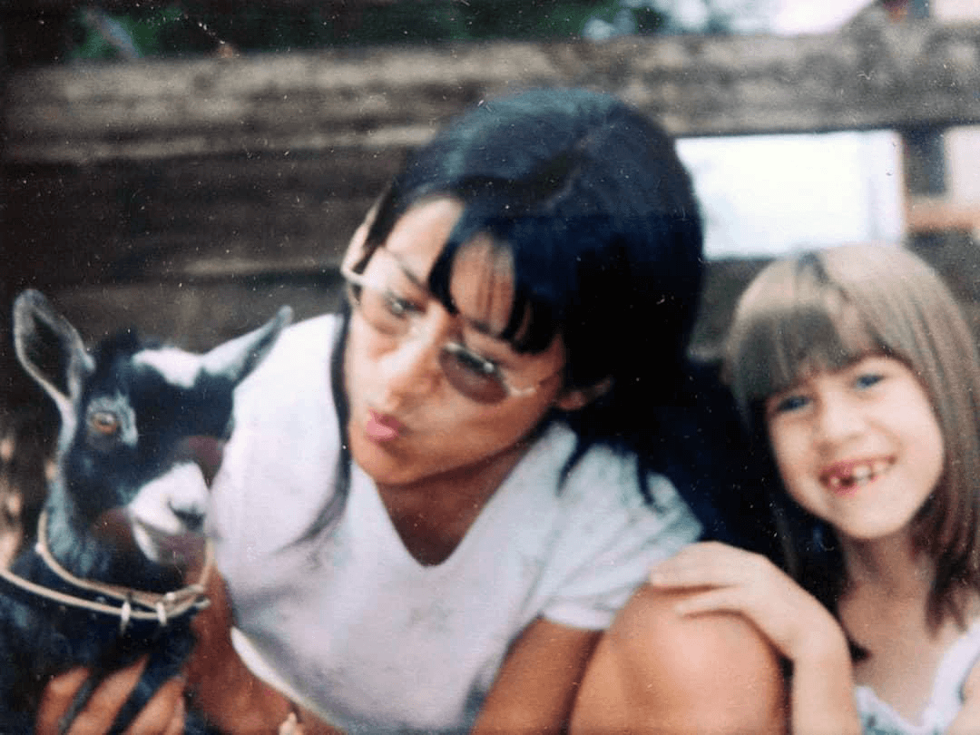 A woman with black hair and glasses, and a young girl with brown hair, smiling together outdoors with a small goat, which the woman is holding.