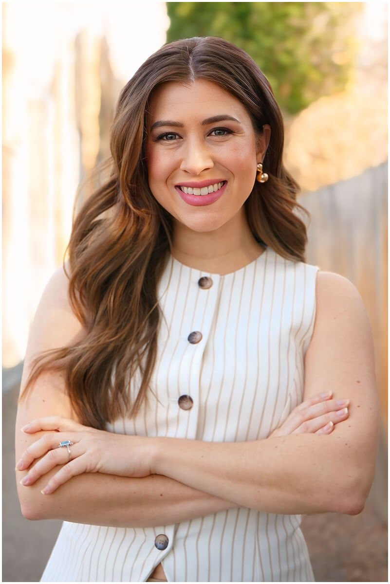 A smiling woman with long, wavy brown hair wearing a sleeveless, white vertically striped top. She has gold earrings and a ring on her left hand. She is outdoors with blurred background trees and sunlight.