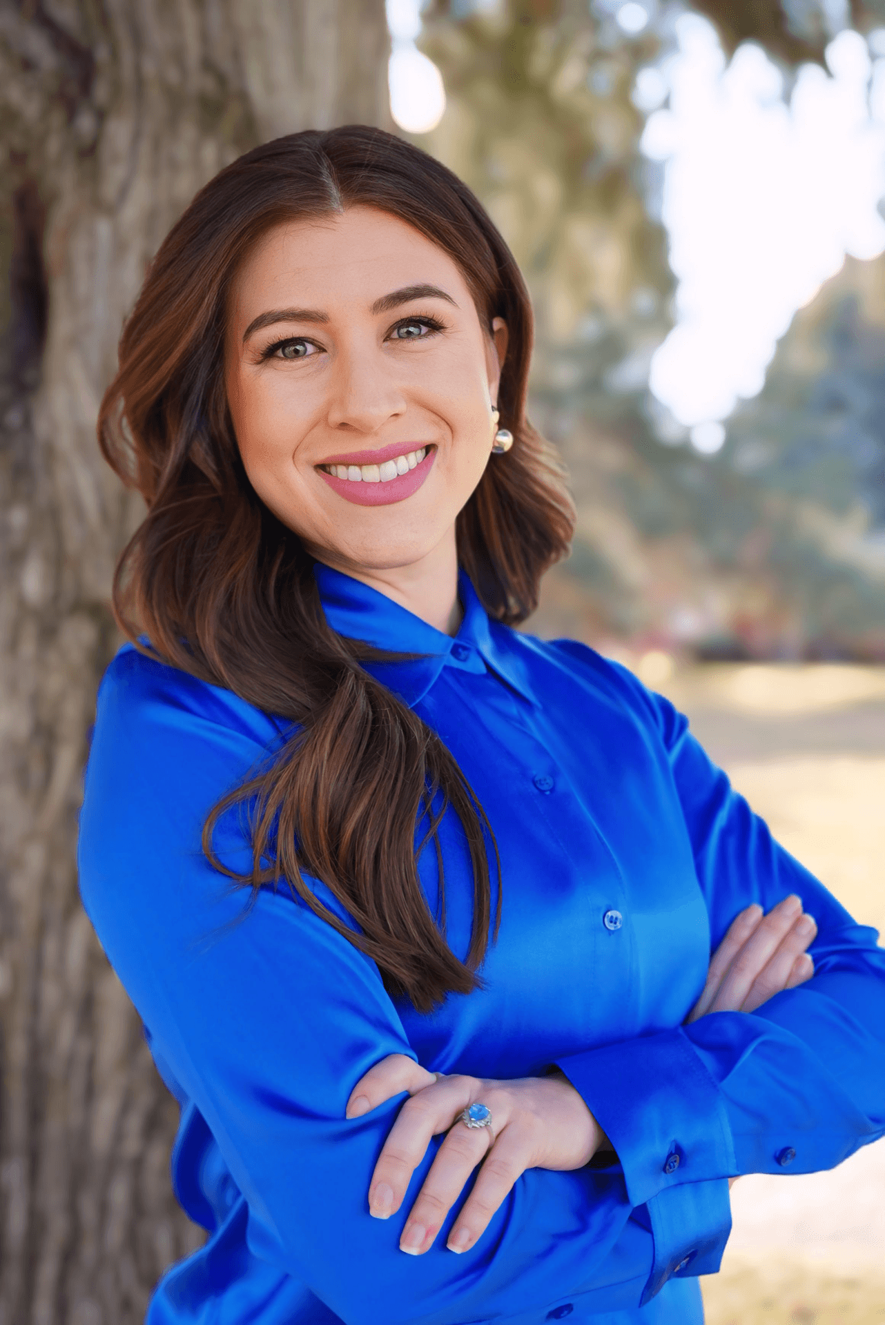 A woman with long brown hair and blue eyes smiling, standing outdoors near a large tree, wearing a bright blue shirt with arms crossed.