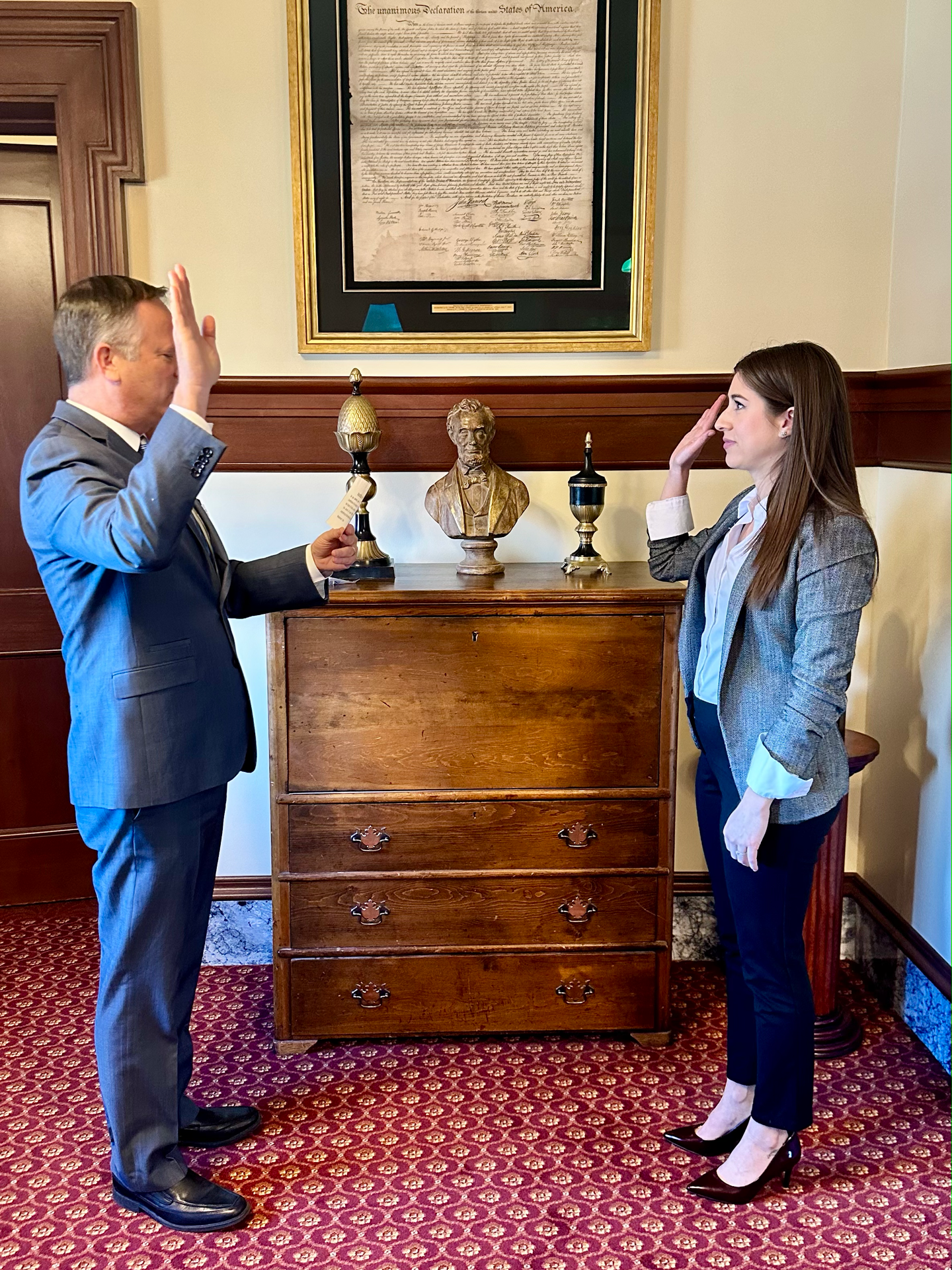 Two individuals, a man and a woman, standing in a room with wood furniture and a red patterned carpet, facing each other with right hands raised in a salute. Behind them is a wooden cabinet with bust and lamp figurines, and a framed document of the Declaration of Independence hanging on the wall.