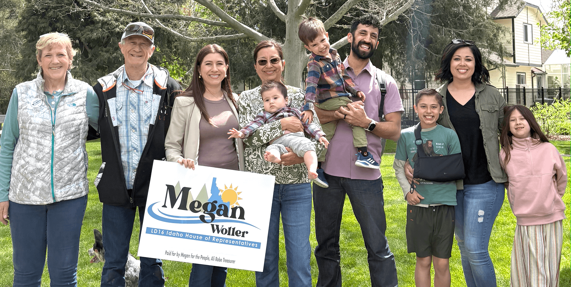 Group of diverse people standing outdoors on grass, smiling at the camera. Some are holding children, including a woman with tree branches in the background. One person holds a sign that reads 'Megan Woller LD16 Idaho House of Representatives,' and a dog is partially visible on the left.