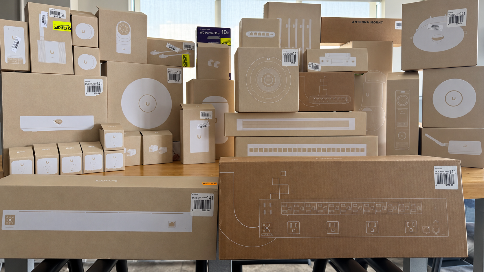 Multiple boxes of various sizes on a table, primarily brown cardboard with white outline drawings of electronic equipment and labels, including a power strip, networking switch, and other electrical components, with some packaging featuring barcodes and price tags.
