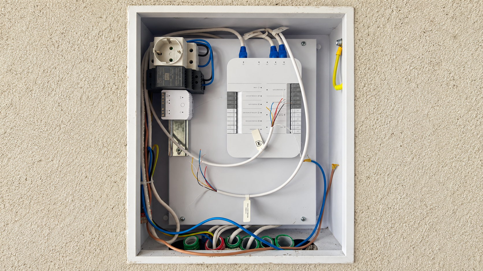 An electrical control panel with wiring, switches, and circuit components mounted in a white metal box on a beige wall.