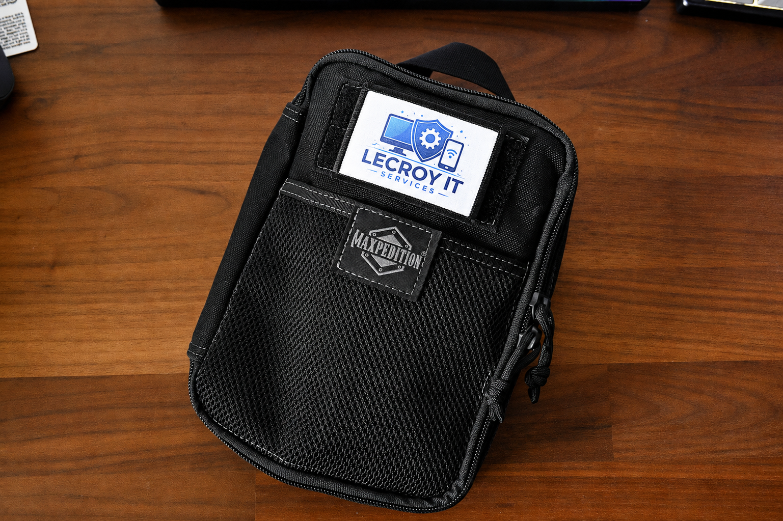 Black Maxpedition tactical pack on a wooden desk with a logo patch and a label that reads 'LECROY IT SERVICES'.