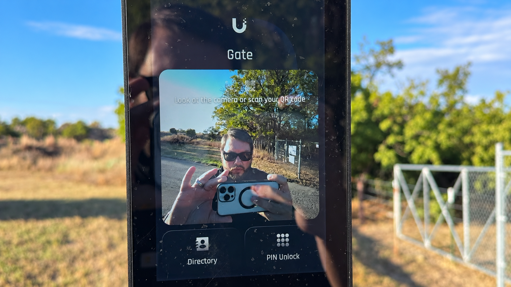 Reflection of a man in a smartphone screen, taking a photo outdoors with trees and a fence in the background, wearing sunglasses and holding a camera.