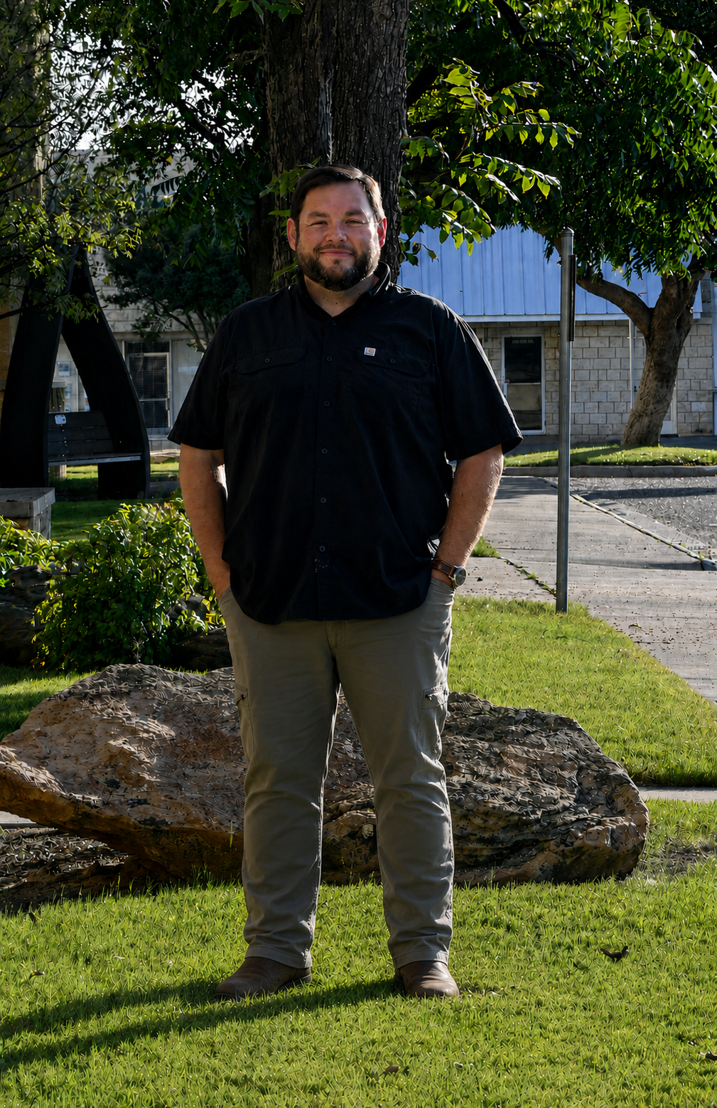 A man stands outdoors on a grass lawn, leaning against a tree, smiling, with sunlight illuminating his face. He is dressed in a black short-sleeved button-up shirt, tan cargo pants, and brown shoes, with a watch on his left wrist.