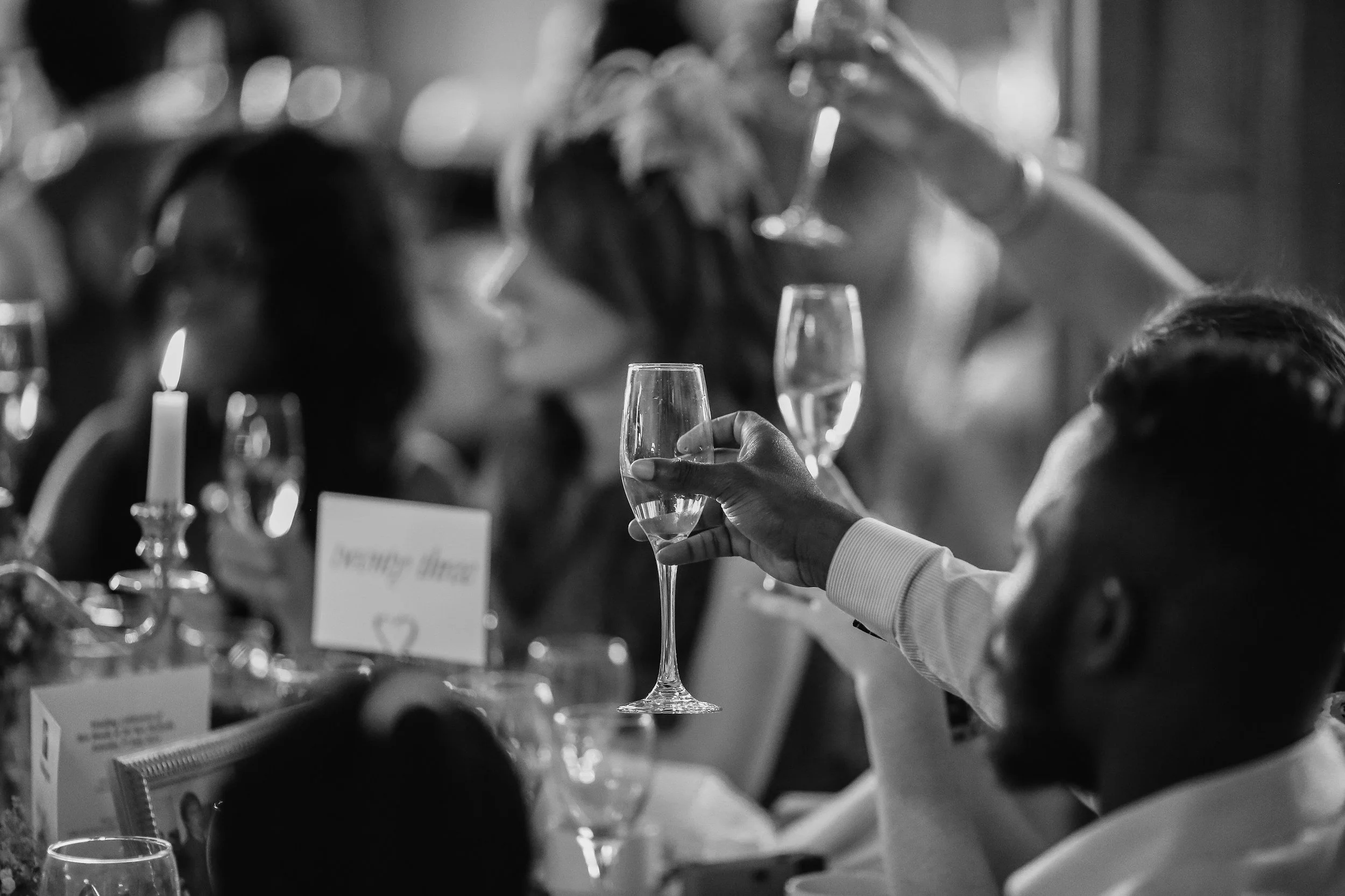 A person holding a champagne flute at a social event or celebration, with blurred guests and lit candles in the background.