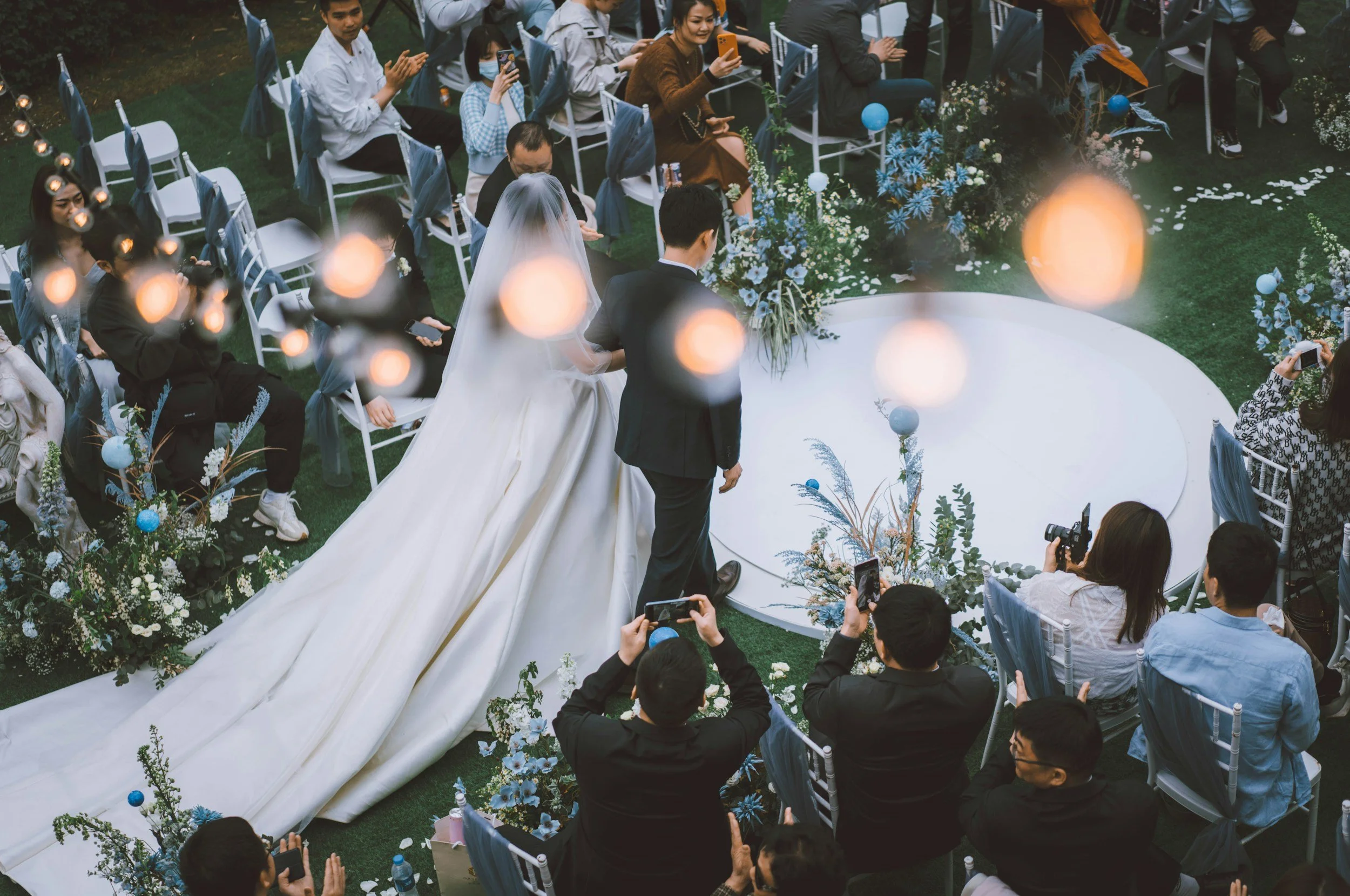 A wedding ceremony outdoors with the bride and groom walking down the aisle, surrounded by guests seated in white chairs. The bride wears a white gown with a long train, and the groom wears a dark suit. The area is decorated with flowers and blue ornaments, with guests taking photos and clapping.