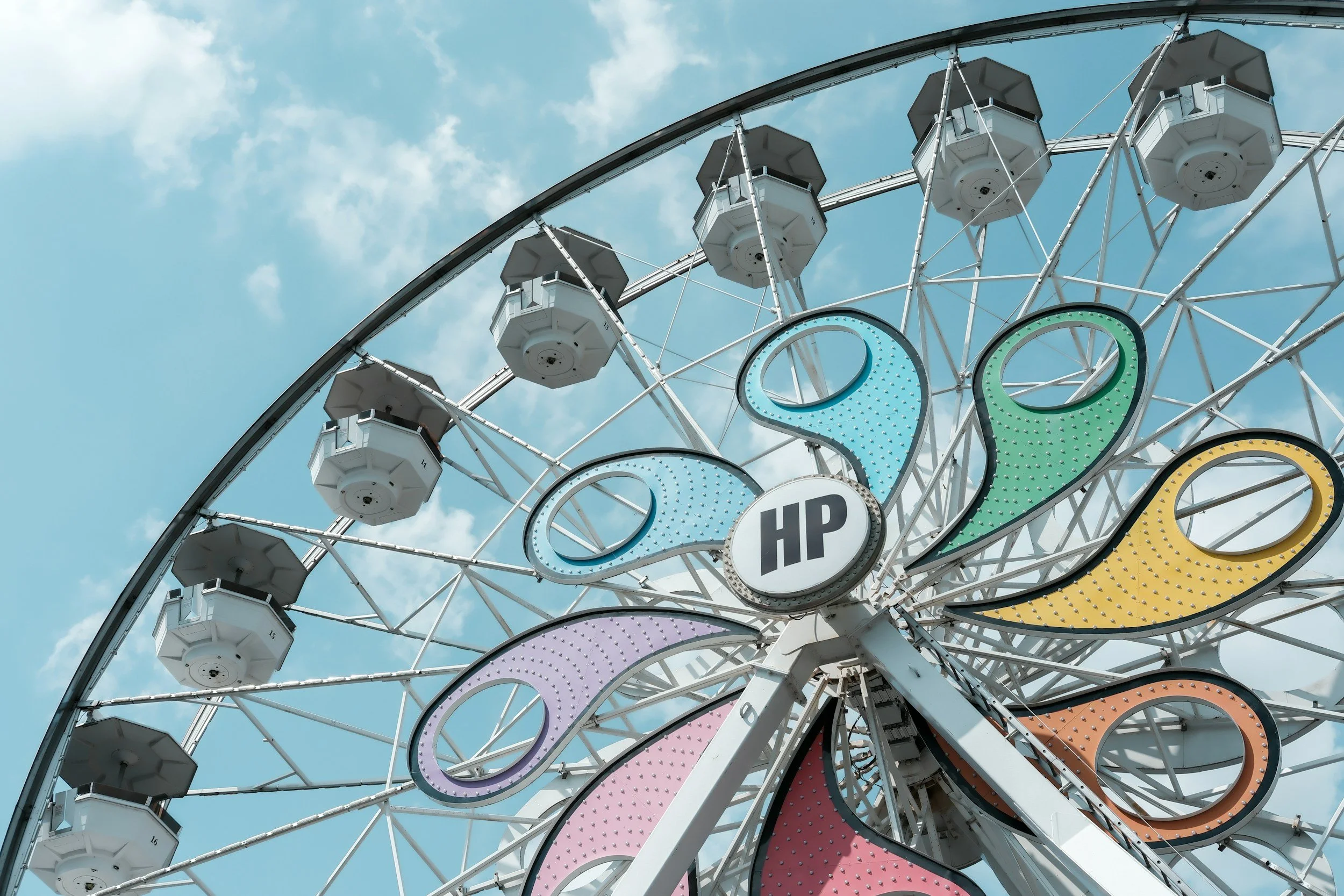 Close-up of a colorful Ferris wheel against a blue sky with clouds, featuring white gondolas and lit-up decorative panels.