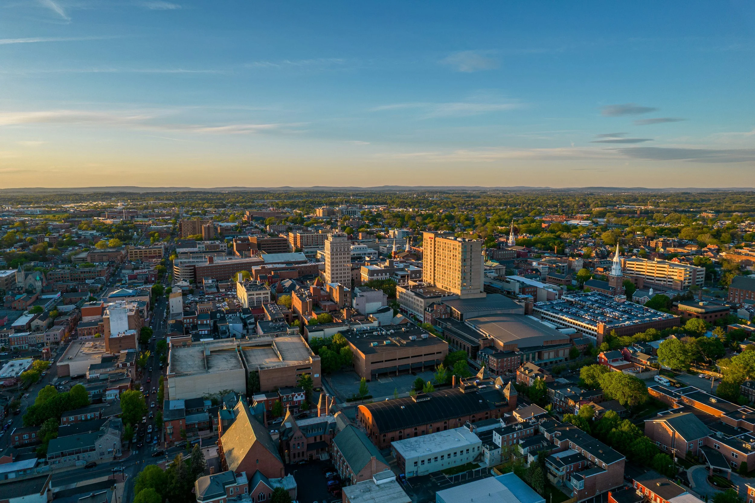An aerial view of a cityscape during sunset with a mix of mid-rise and low-rise buildings, green trees, and streets, under a partly cloudy sky.