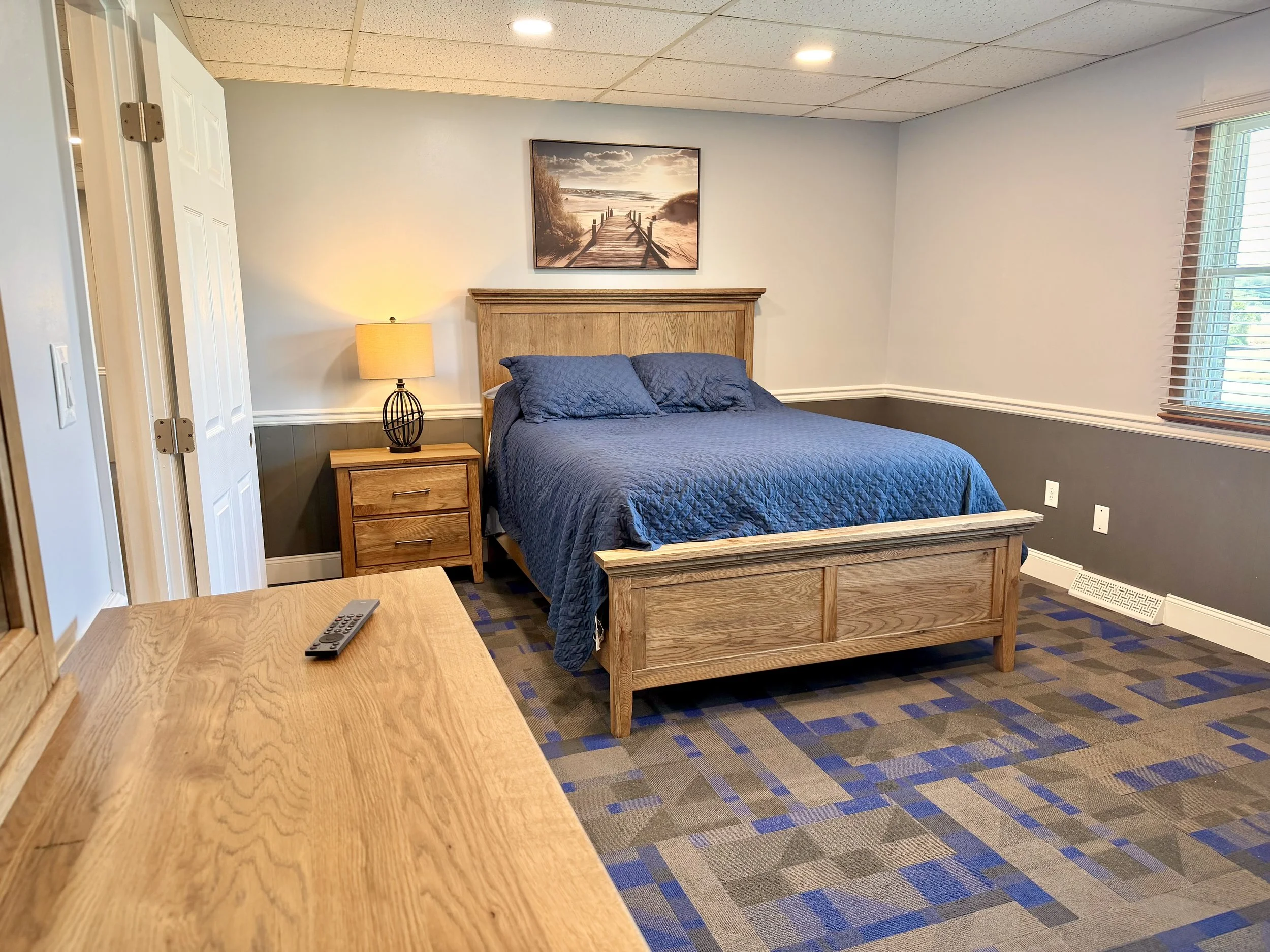 A bedroom with a wooden bed, blue quilt, and pillows, a bedside table with a lamp, a painting of a beach scene above the bed, a window with blinds, and a patterned carpet.