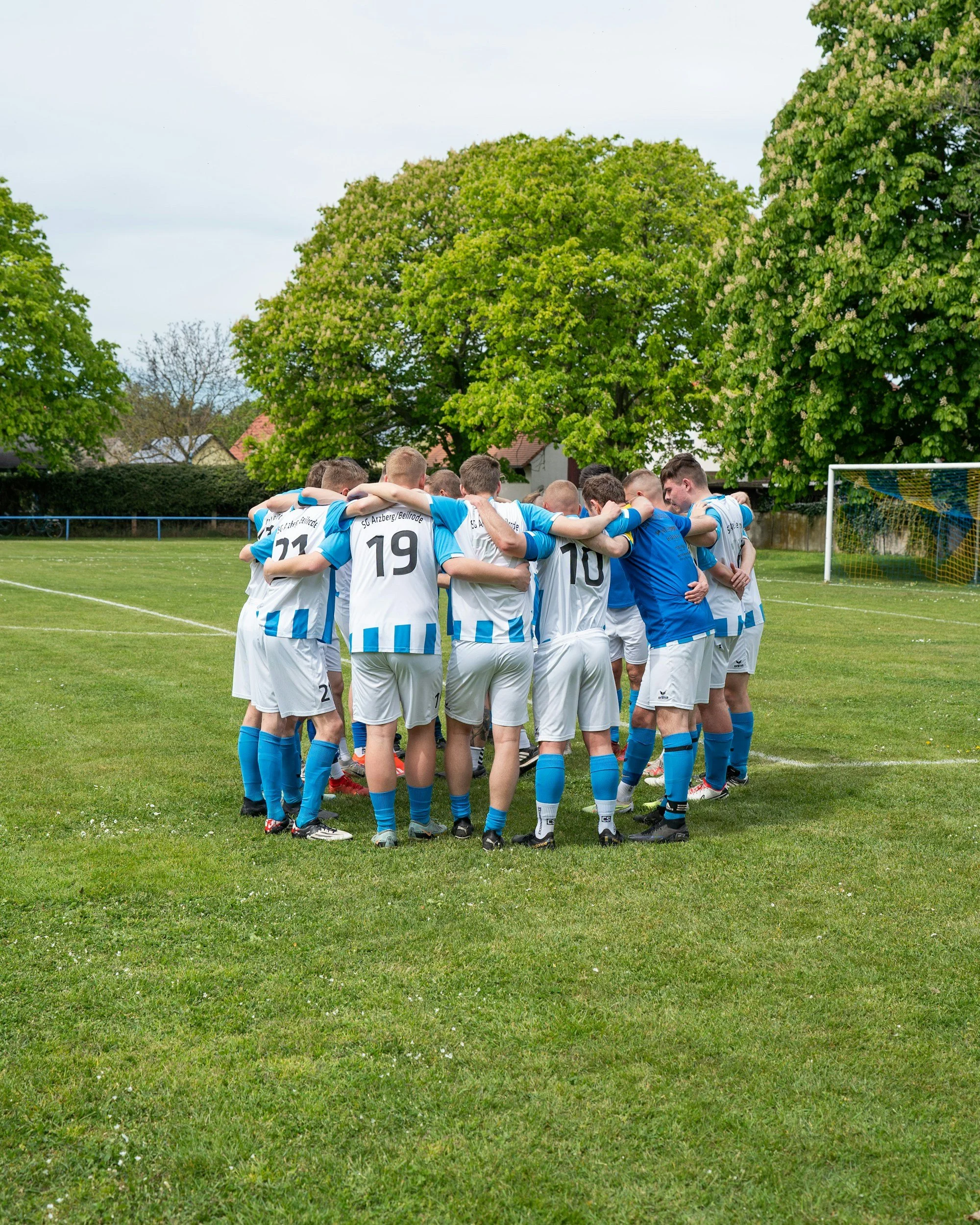 Soccer team huddling on a grassy field before a game, with large green trees and a blue sky in the background.