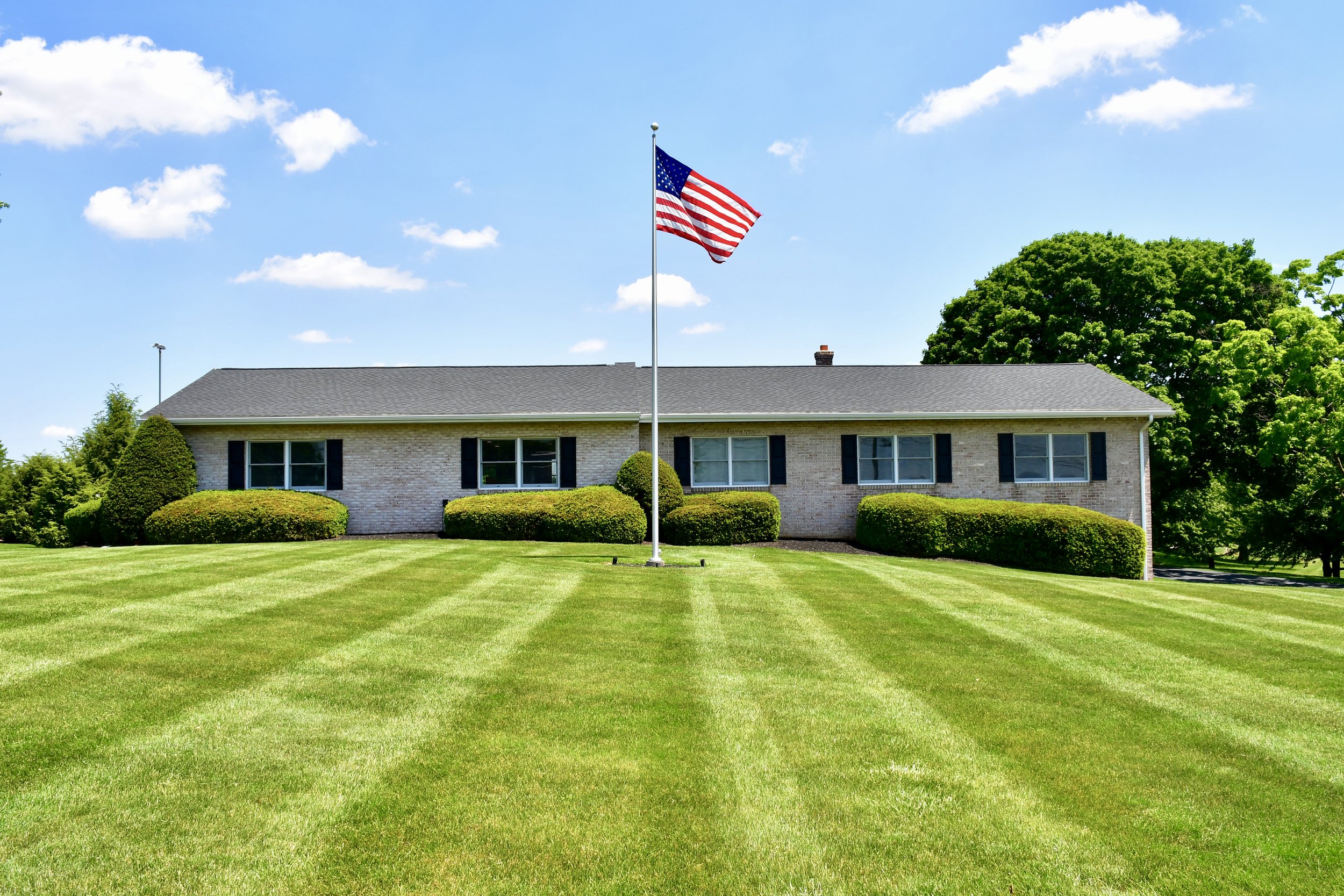 A house with a gray roof, brick walls, and black shutters, surrounded by neatly trimmed bushes and a green lawn. An American flag is flying on a flagpole in the front yard, with a bright blue sky and some white clouds in the background.