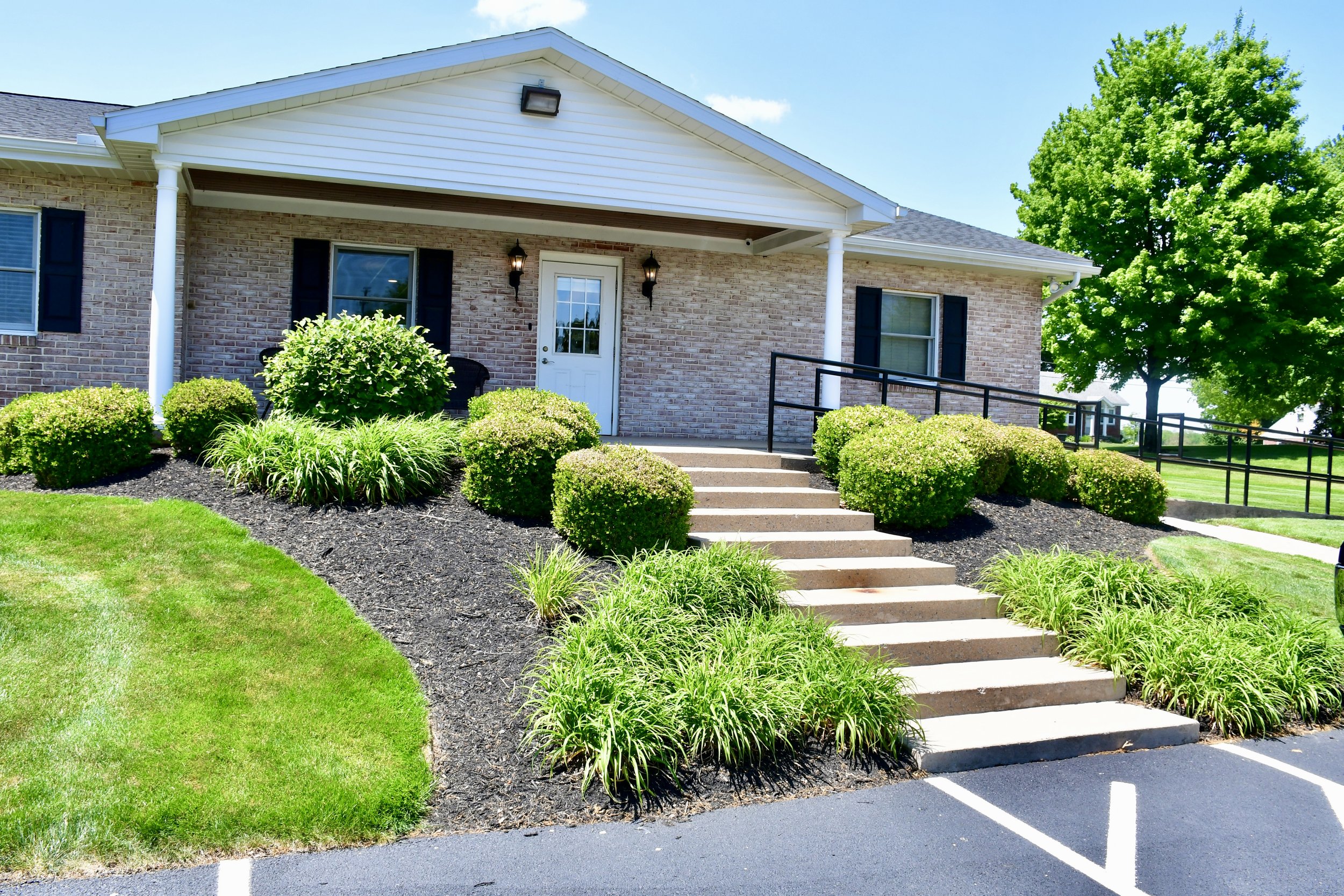 Front view of a brick house with a white gabled roof, surrounded by well-maintained green bushes, a lush tree, and a small porch with stairs leading to a white door.