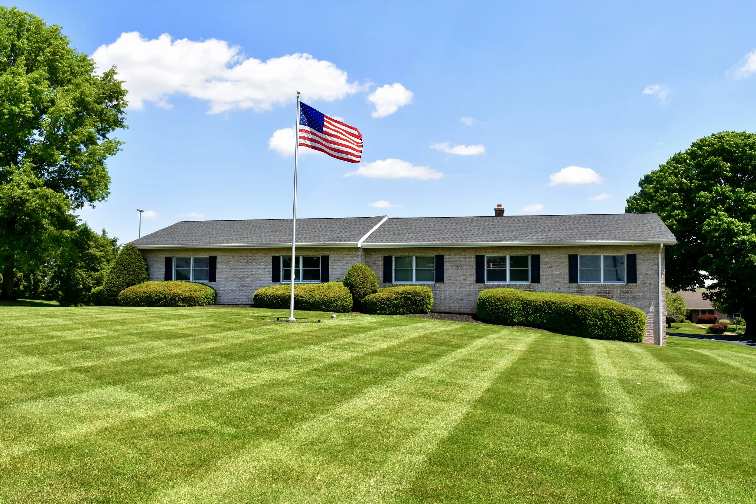 American flag flying on a flagpole in front of a house with a well-manicured lawn and trees under a blue sky with scattered white clouds.