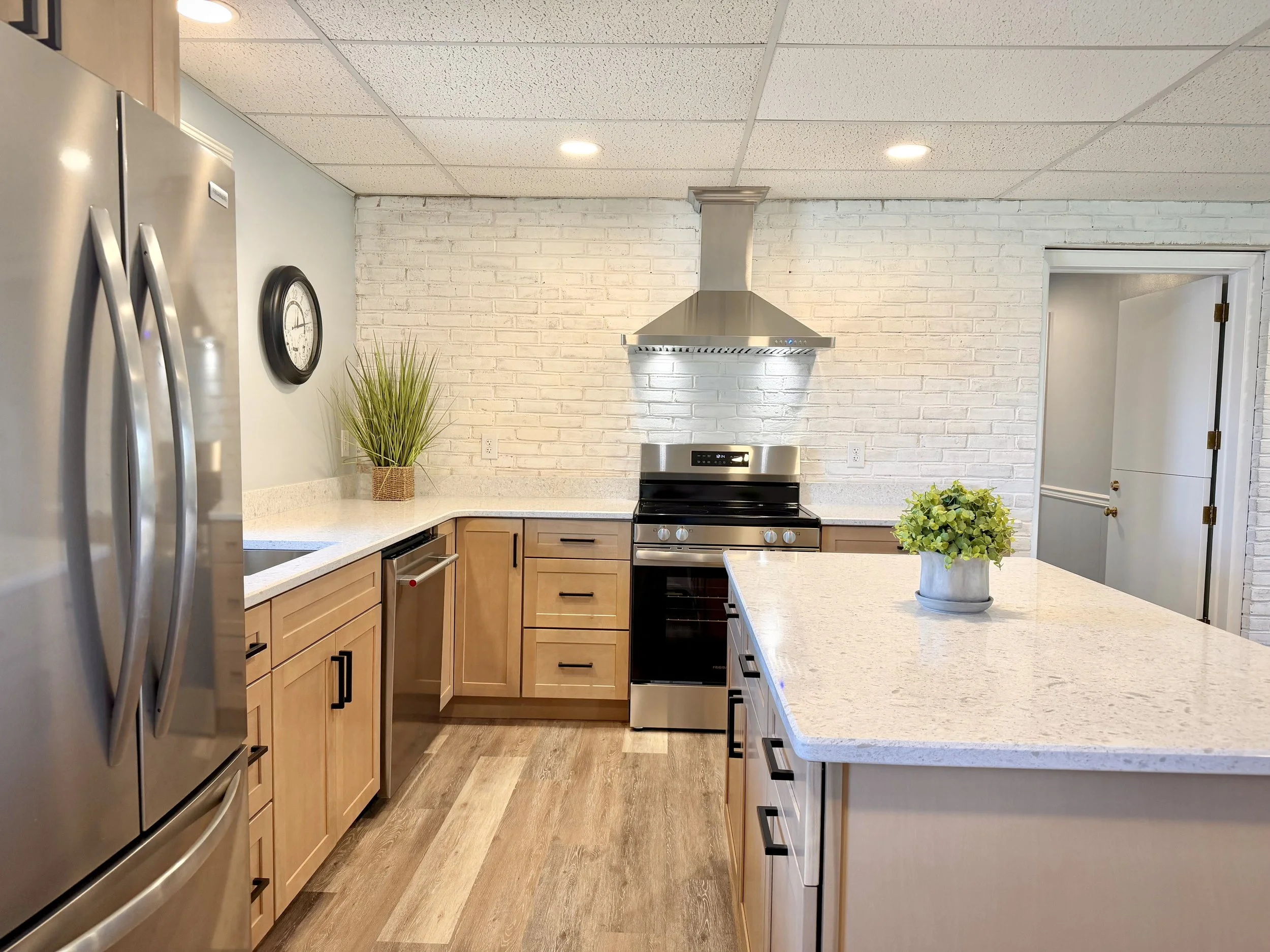 Modern kitchen with white brick backsplash, wooden cabinetry, stainless steel appliances including a refrigerator, oven, and range hood. A potted plant on the countertop and a wall clock on the wall.