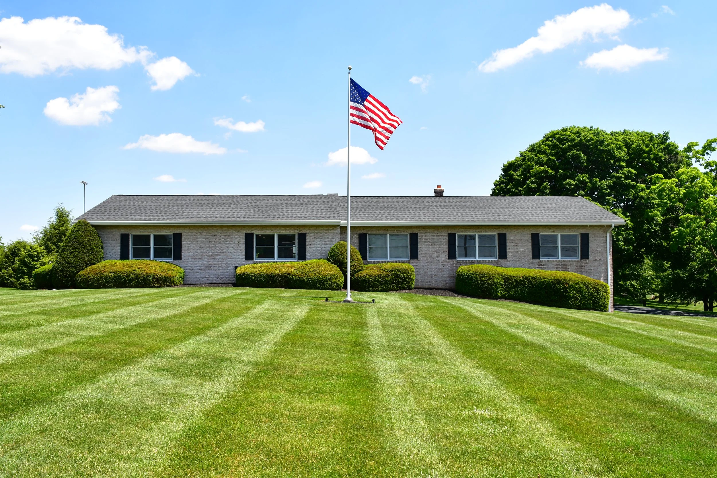 A house with a neatly maintained lawn, American flag on a tall flagpole in front, and large green trees behind under a bright blue sky with scattered white clouds.