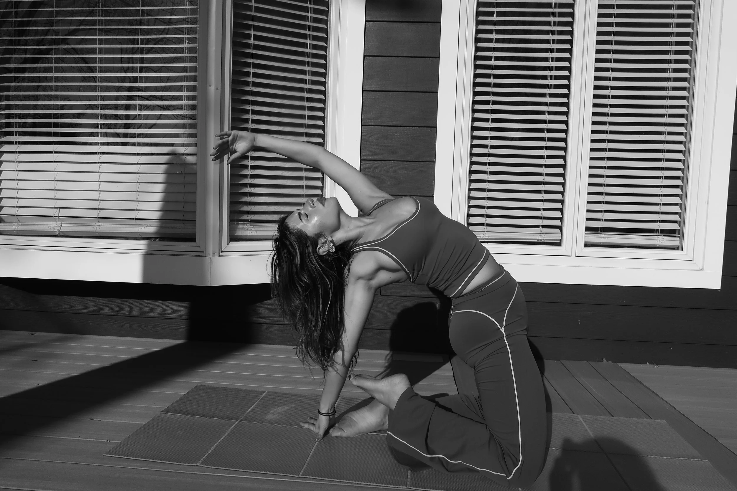A woman practicing yoga outdoors on a mat in front of a building with wooden and glass windows, performing a side bend pose with one arm reaching over her head and the other supporting her body.