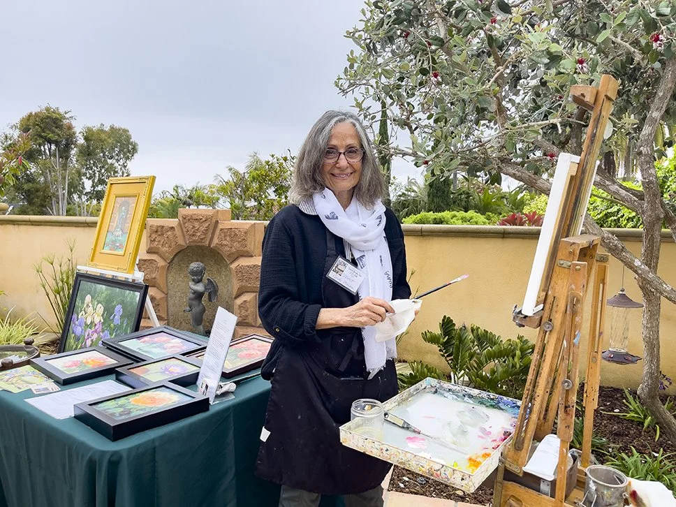 A female artist smiles at the camera and stands in front of her canvas while holding a paint brush.