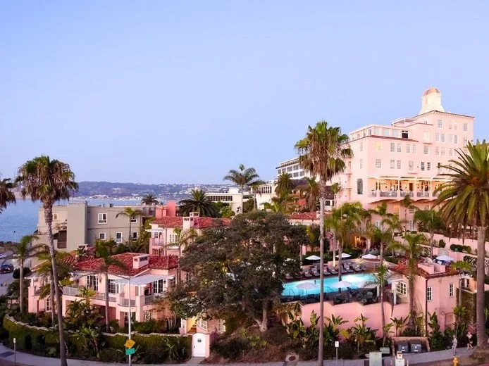view of La Jolla's La Valencia Hotel and surrounding businesses. A pink hotel surrounded by palm trees.