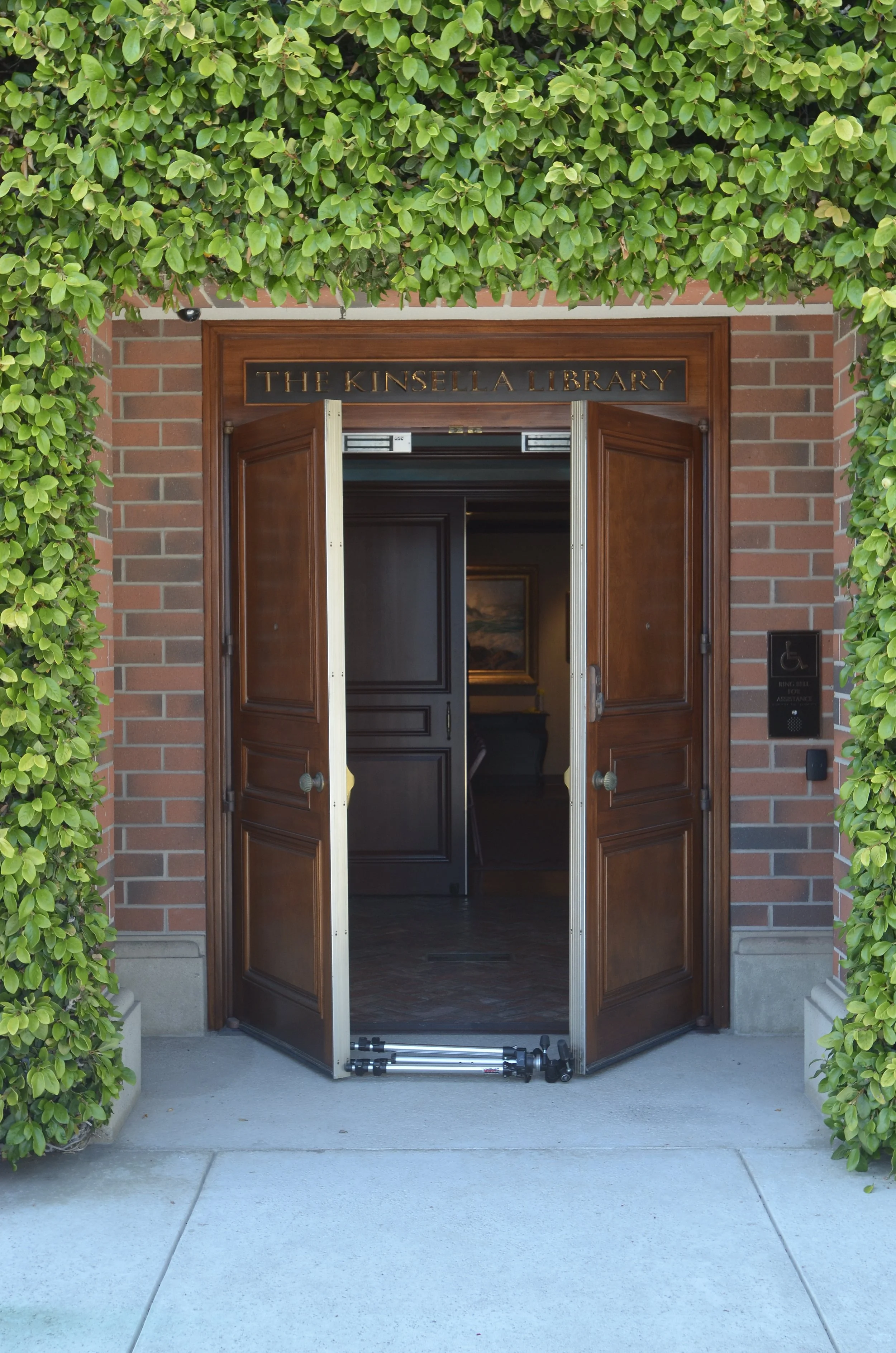 brick framed door propped open, surrounded by ficus. The sign above the book reads: The Kinsella Library
