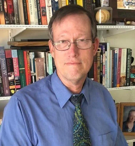 man with glasses and blue shirt and green paisley tie in front of a bookshelf with a baseball
