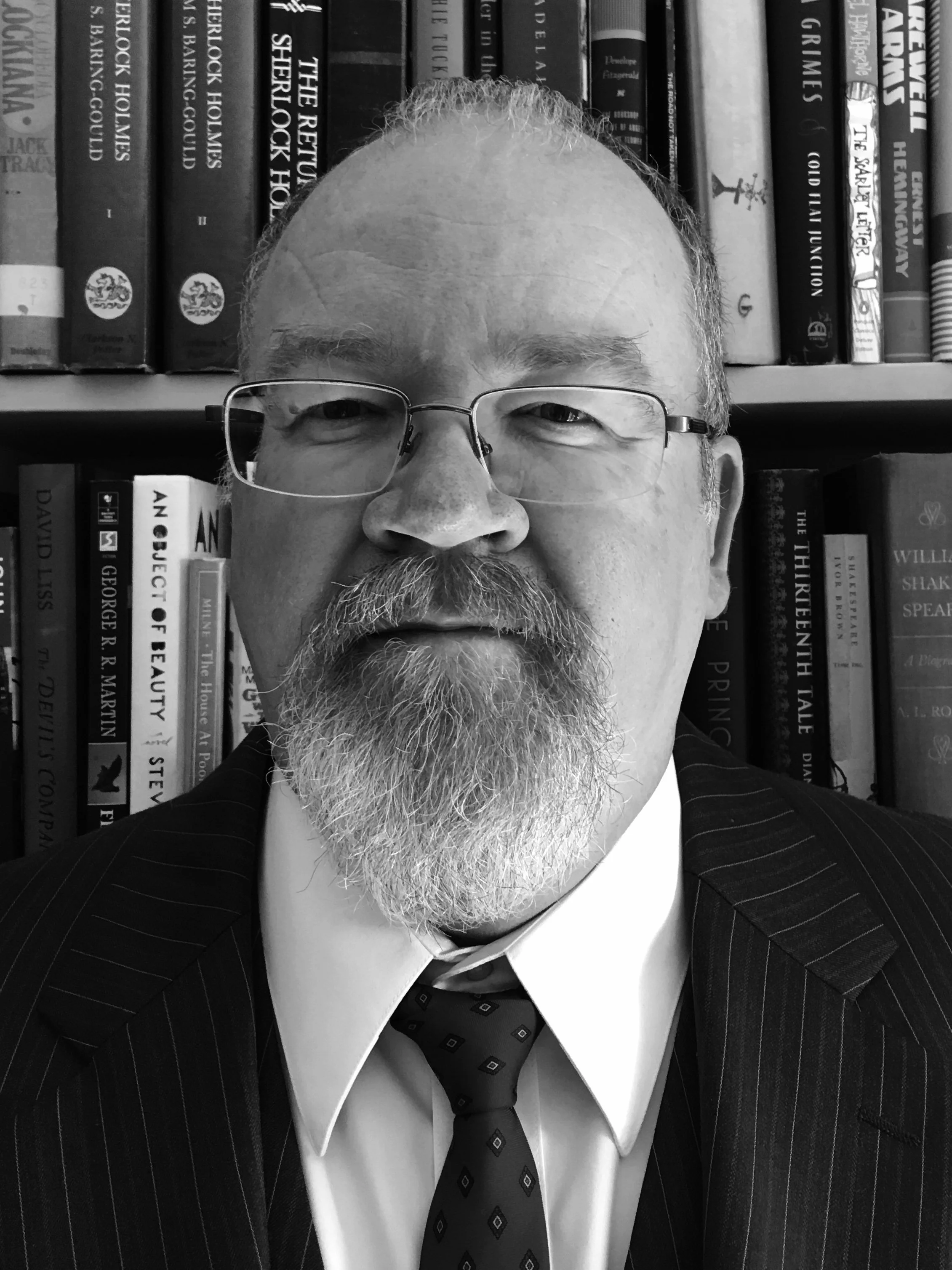 black and white photo of man with a goatee and glasses in front of a bookshelf