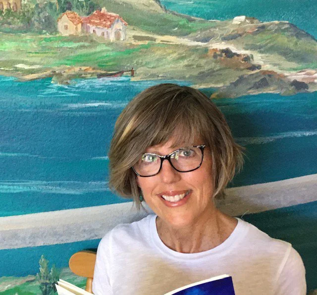 woman reading in front of a landscape painting. Smiling and wearing glasses and white shirt