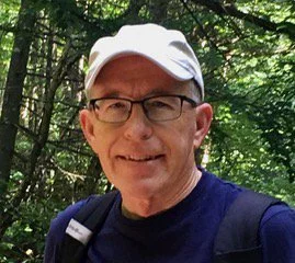 man smiling with hat, glasses and backpack in navy blue shirt