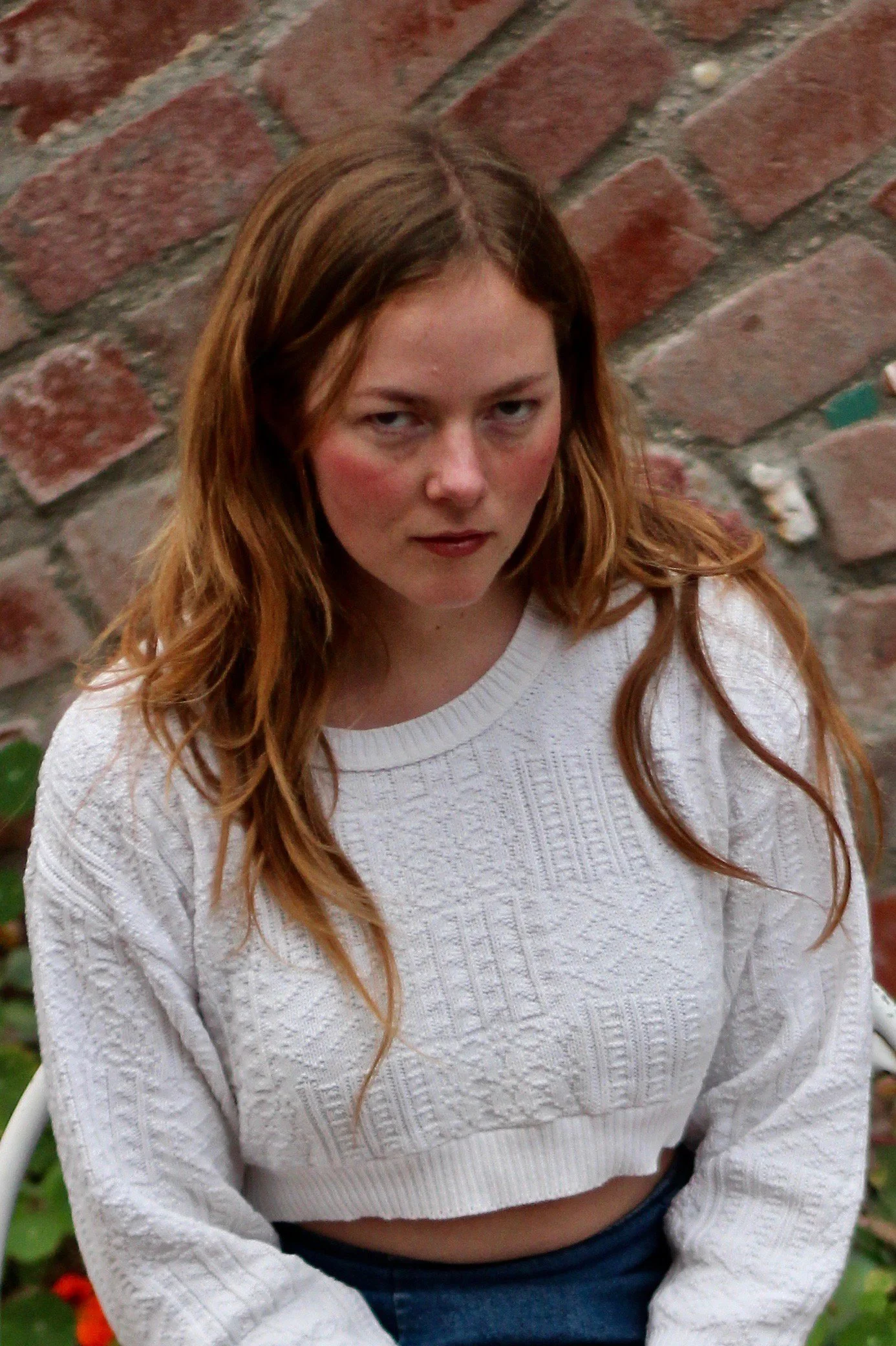 young woman with dark red long hair in a white sweater in front of a brick wall