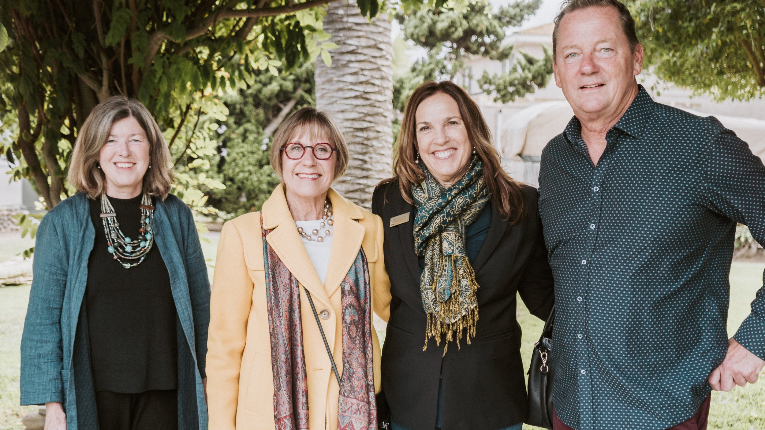 Three people smile at the camera with their arms around each other and a large palm tree is partially seen behind them.