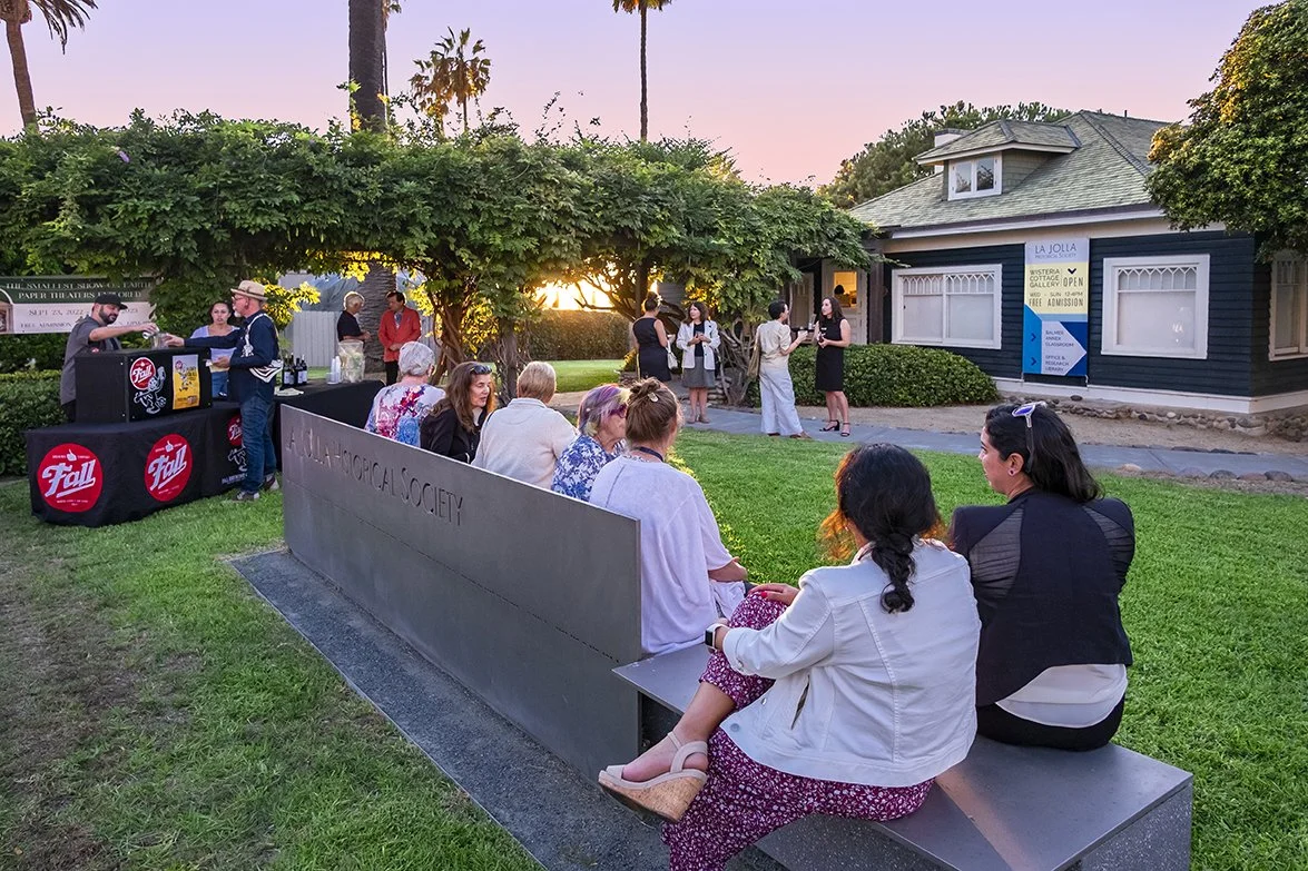 A group of people stand or sit across a long metal bench in front of a historic cottage and vine-covered pergola.