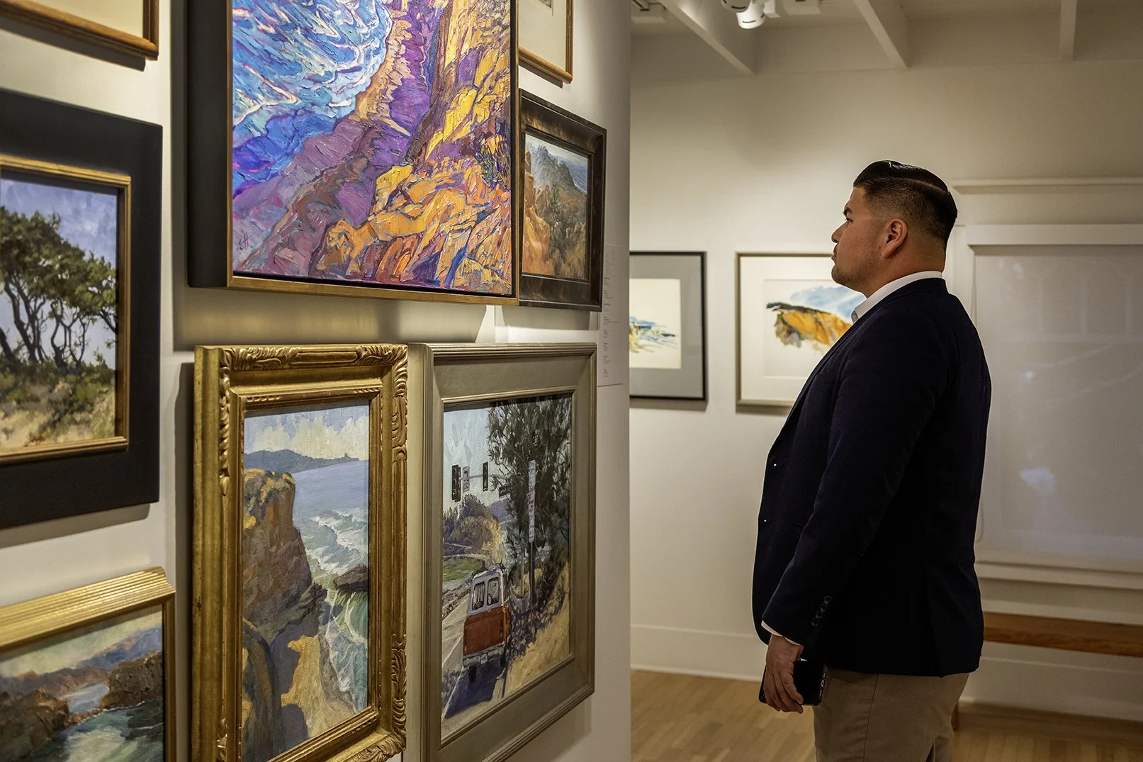 A man in a blue coat stares at a wall covered in colorful landscape paintings.