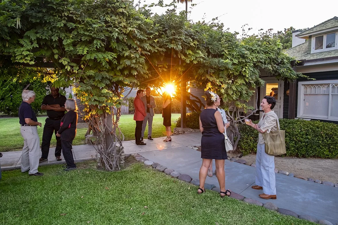 A group of people stand across a lawn, sidewalk and under a vine-covered pergola with the sun setting.