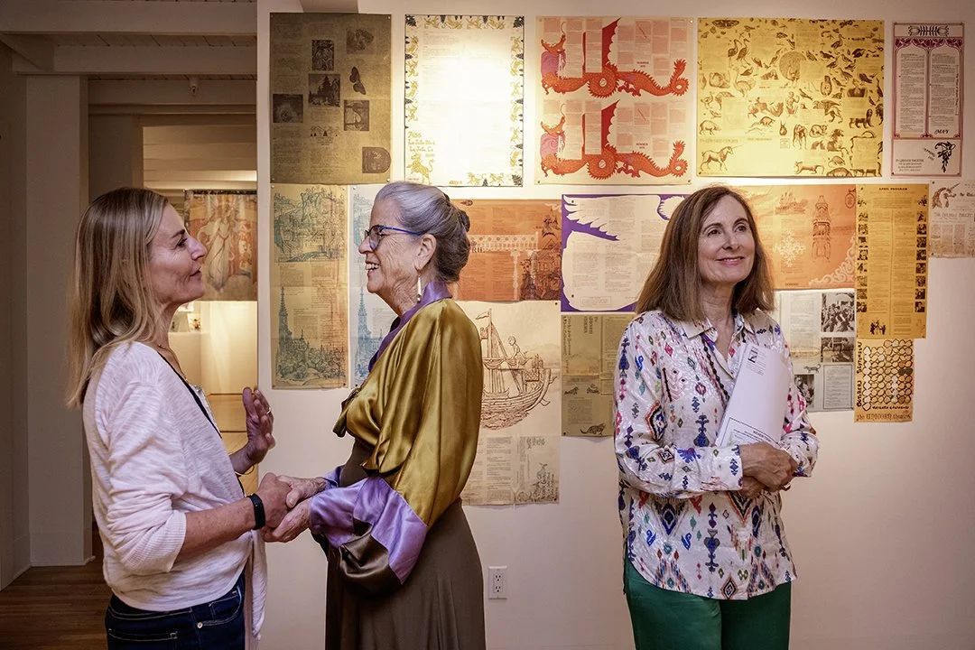 Three women stand together in an art gallery smiling and two holds hands.