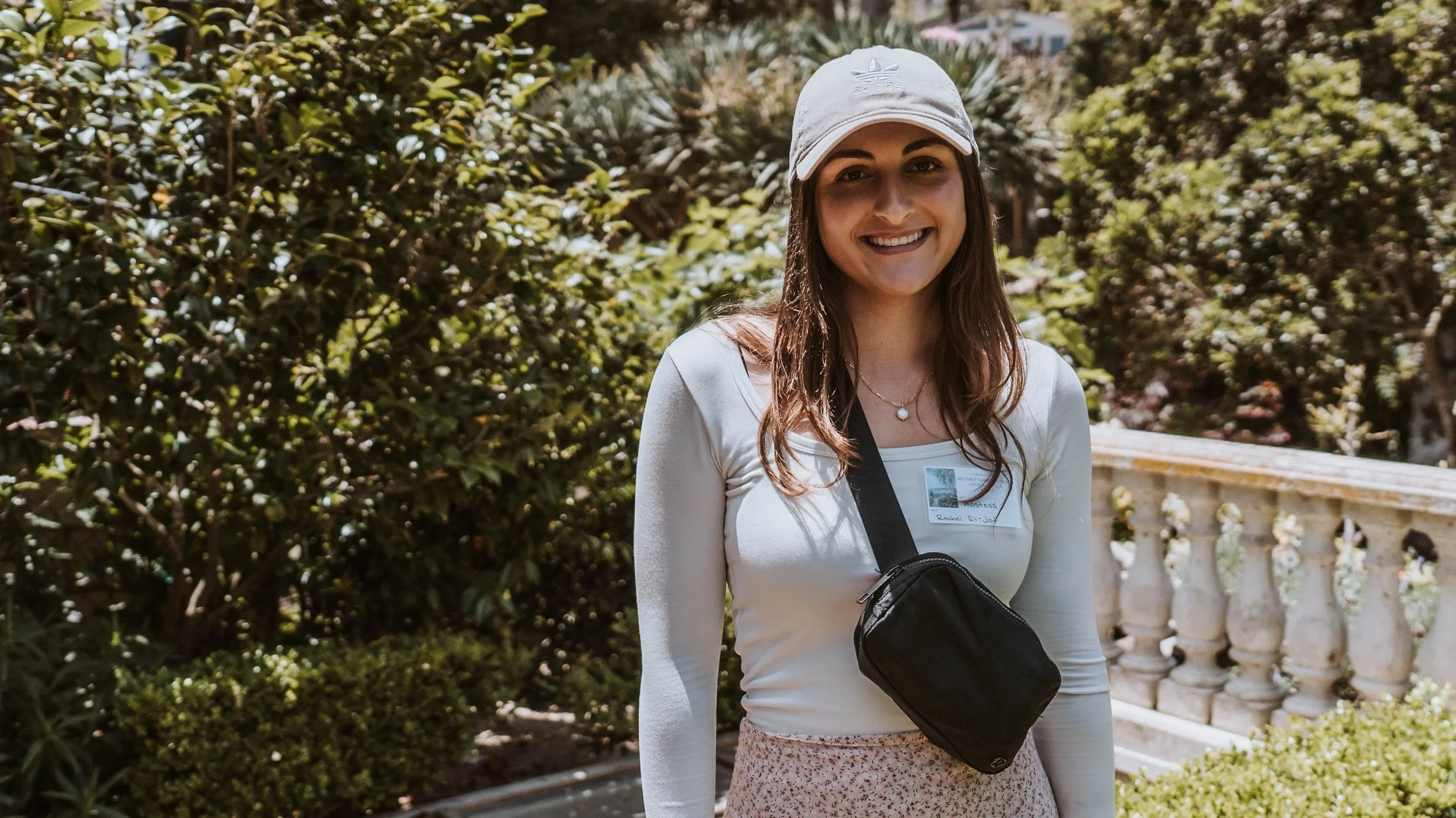 A young woman smiles at the camera while standing in a lush, green garden.