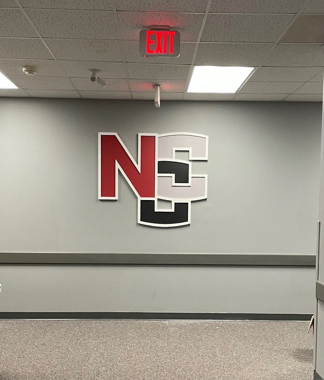 Interior wall with a large NC State logo and an illuminated red 'EXIT' sign above it.