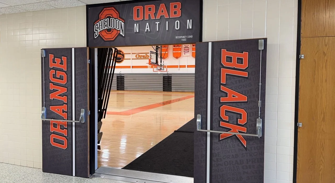 Entrance to a basketball gymnasium at Sheldon High School, with banners reading 'ORAB NATION' and 'ORANGE' and a view of the basketball court inside.