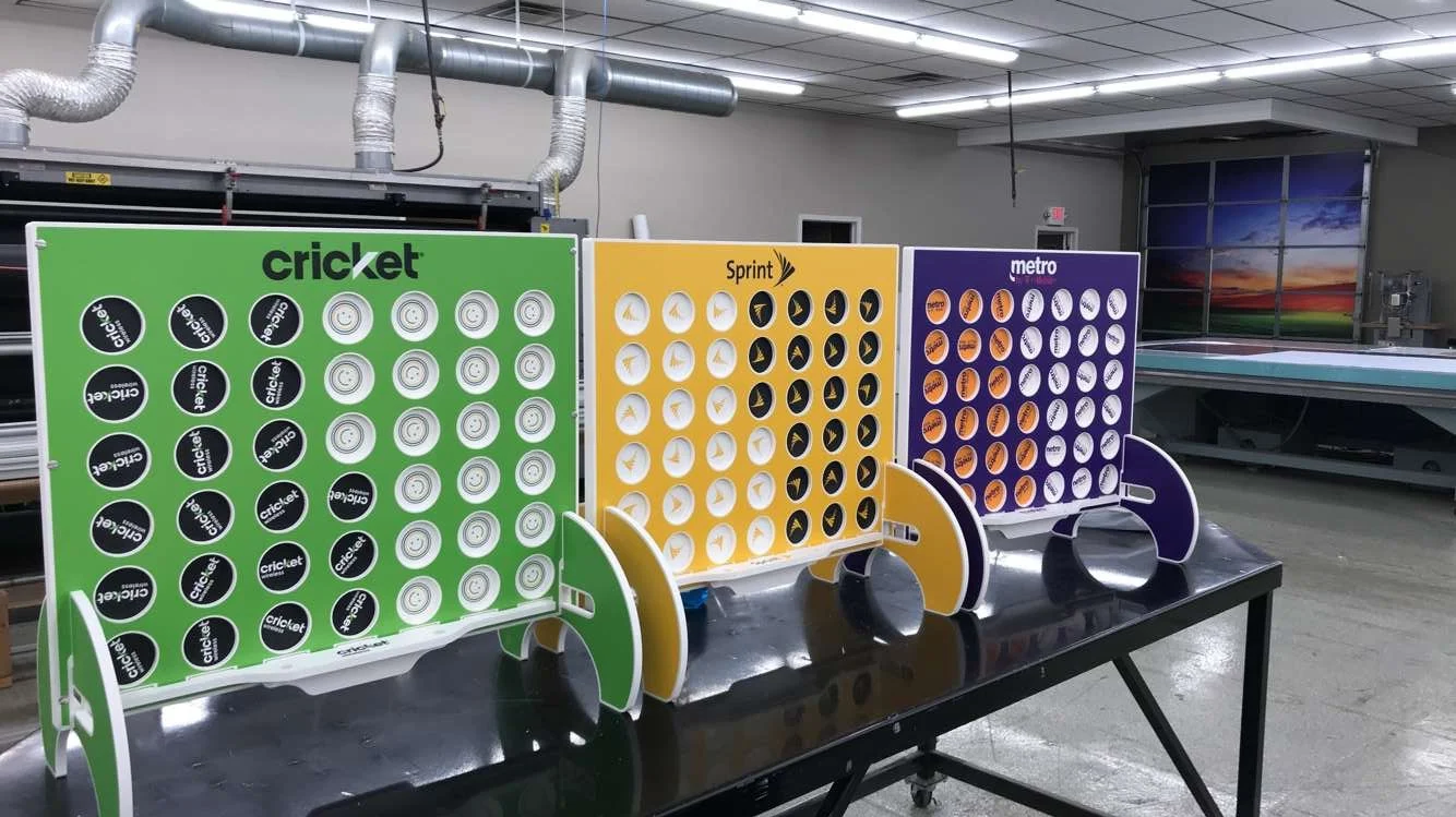 Three vertical wooden board games labeled 'cricket', 'Sprint', and 'metro' with circular holes, fitted into a black table in a room with industrial ceiling and a large window showing a sunset sky.
