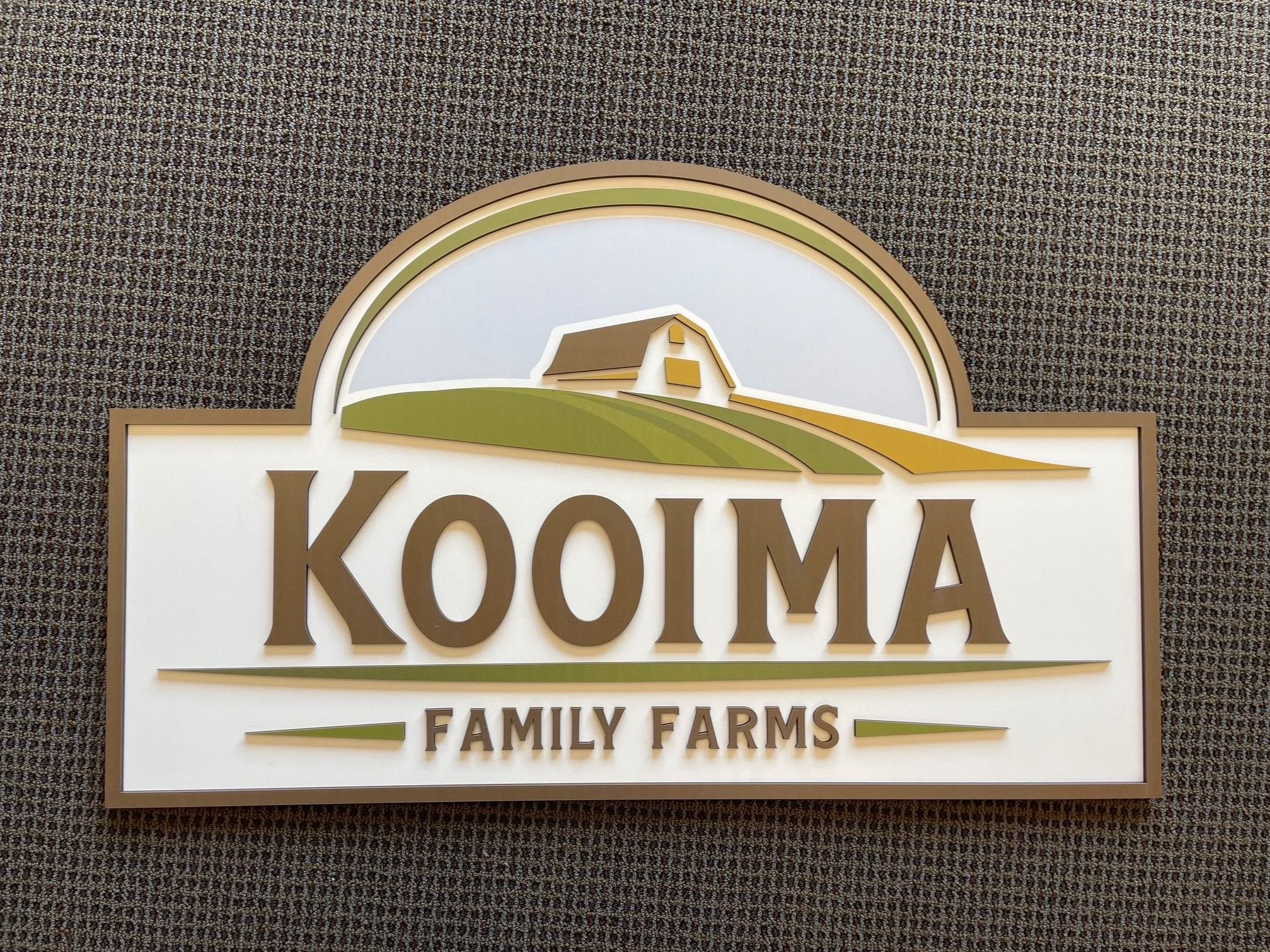 Sign for KOOM Farms featuring a barn illustration and green fields on a textured brown background.