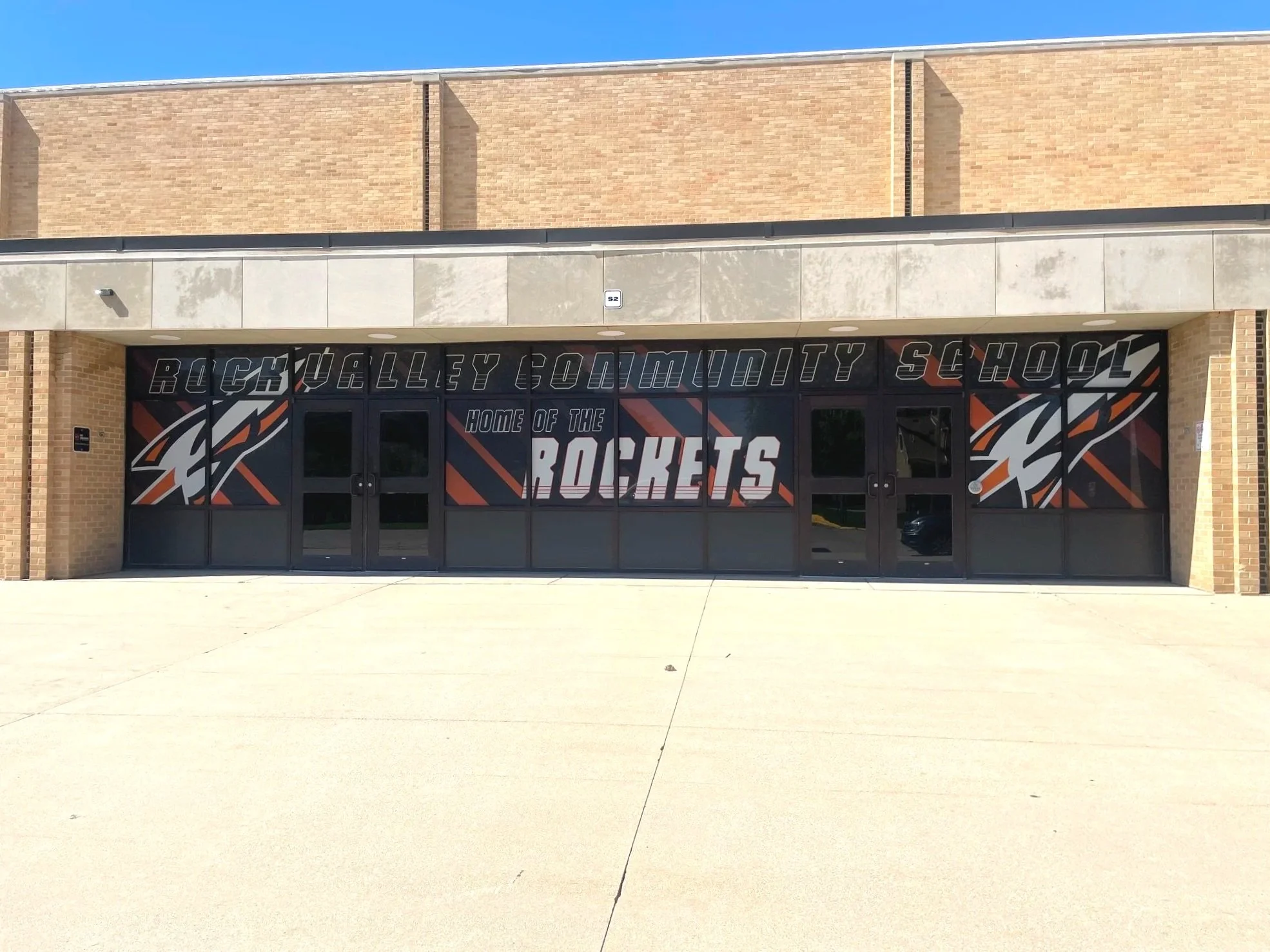 Entrance of Rock Valley Community School with a large sign displaying the school's name and mascot, a rocket, in front of a brick building under a clear blue sky.