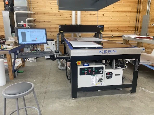 A large CNC cutting machine labeled 'KERN' in a workshop, connected to a computer monitor displaying a CAD design.
