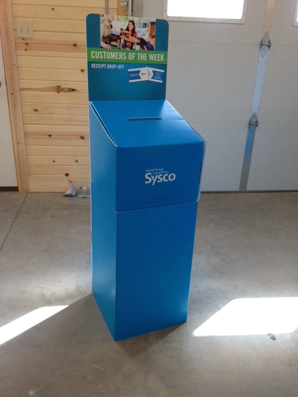 Blue donation drop-off box labeled 'SYSCO' with a sign on top indicating 'Customers of the Week' and 'Receipt Drop-Off'.