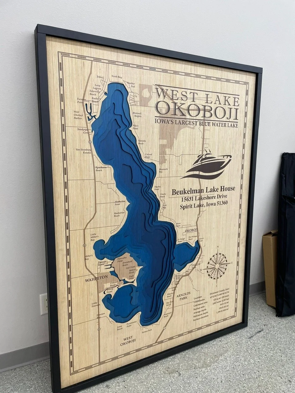 Map of West Lake Okoboji, Iowa, with a blue water body illustration, showing surrounding neighborhoods and landmarks, framed in black, leaning against a wall.